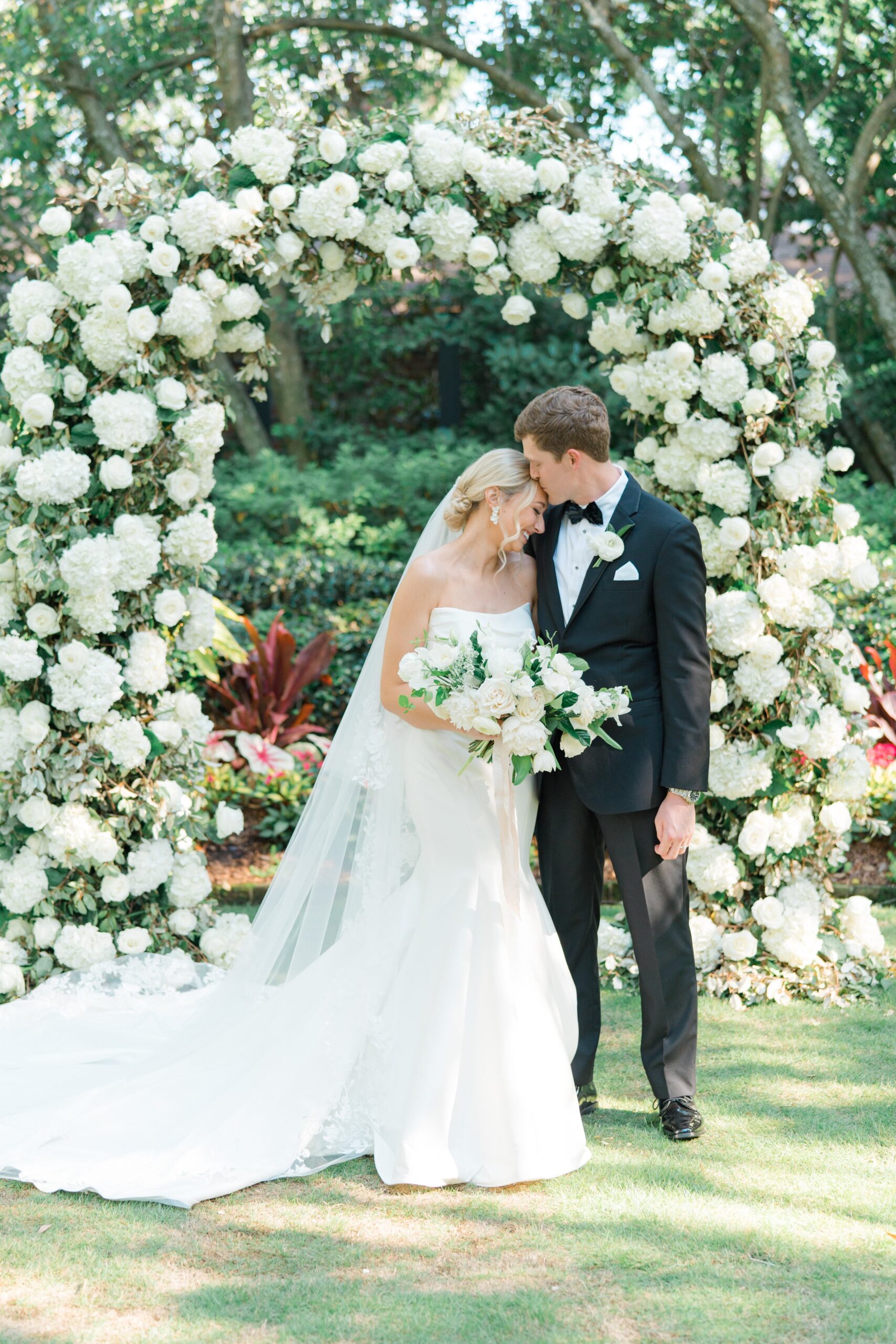 Groom kisses brides forehead under lush white flower arch Governor Thomas Bennett House wedding ceremony. Spring wedding in Charleston