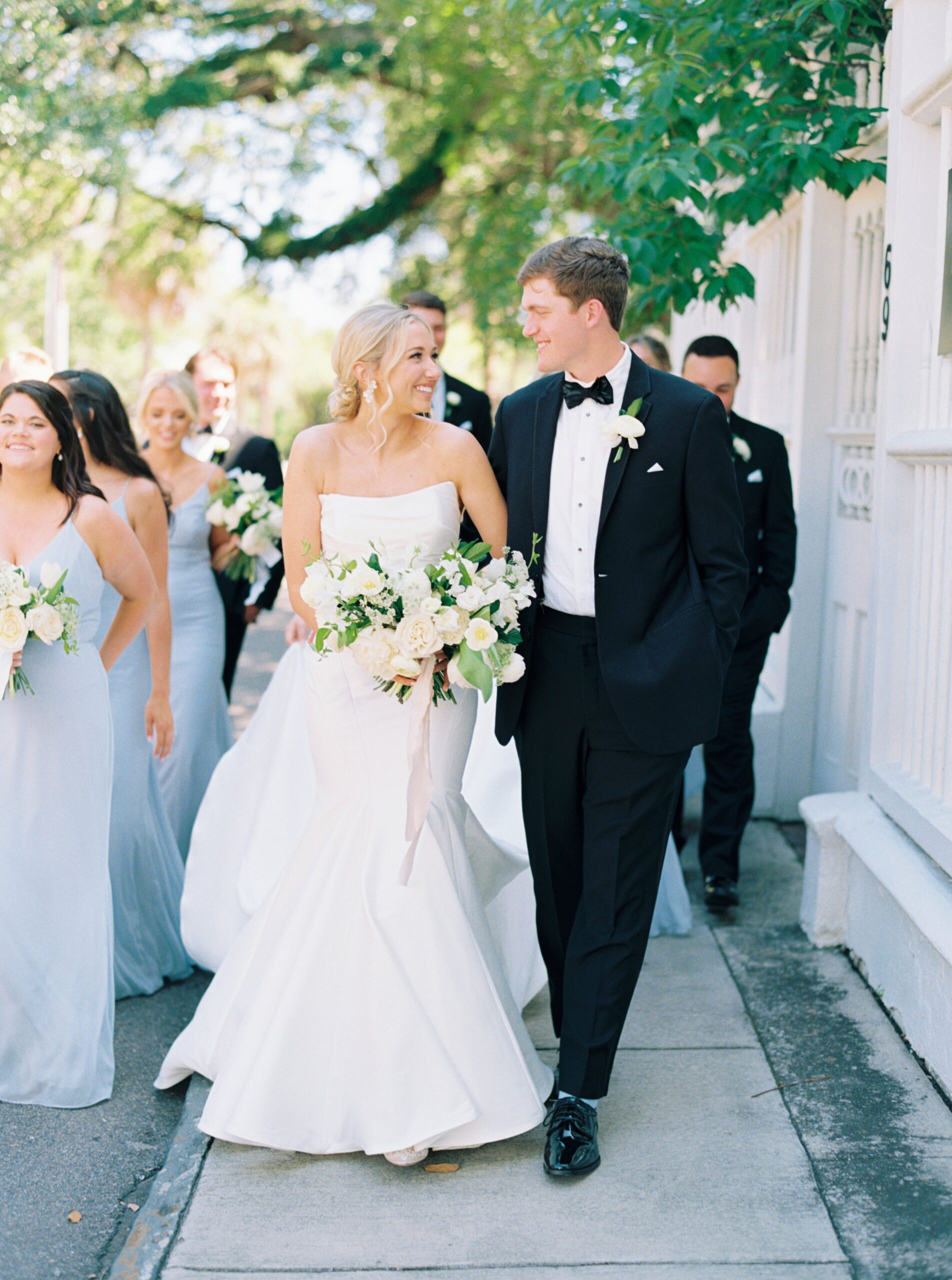 Charleston wedding film photographer. Bride and groom walking on sidewalk with white fence and greenery.