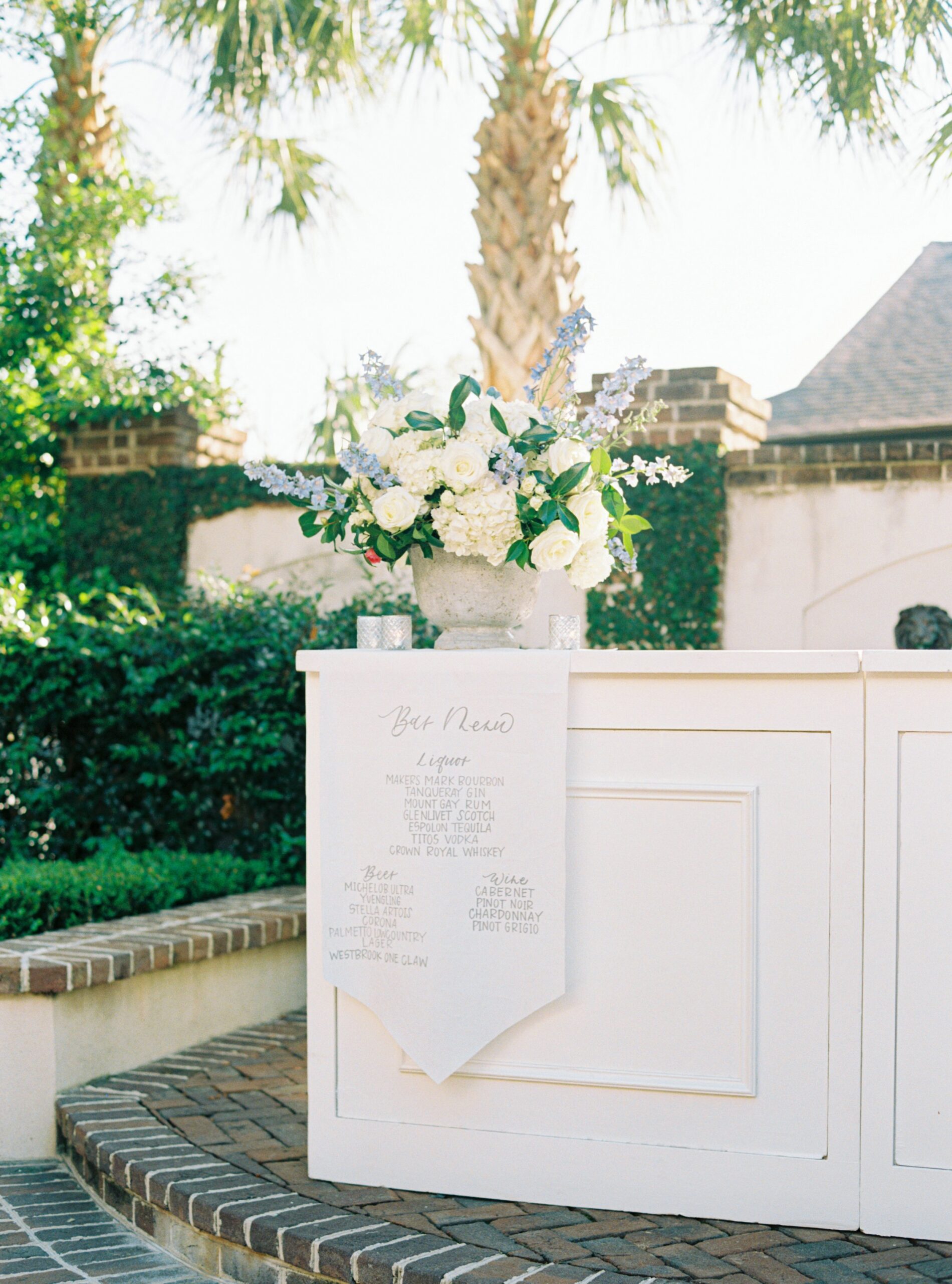 Outdoor wedding reception bar with custom sign. White flowers with a touch of light blue. Palm tree at spring Charleston wedding.