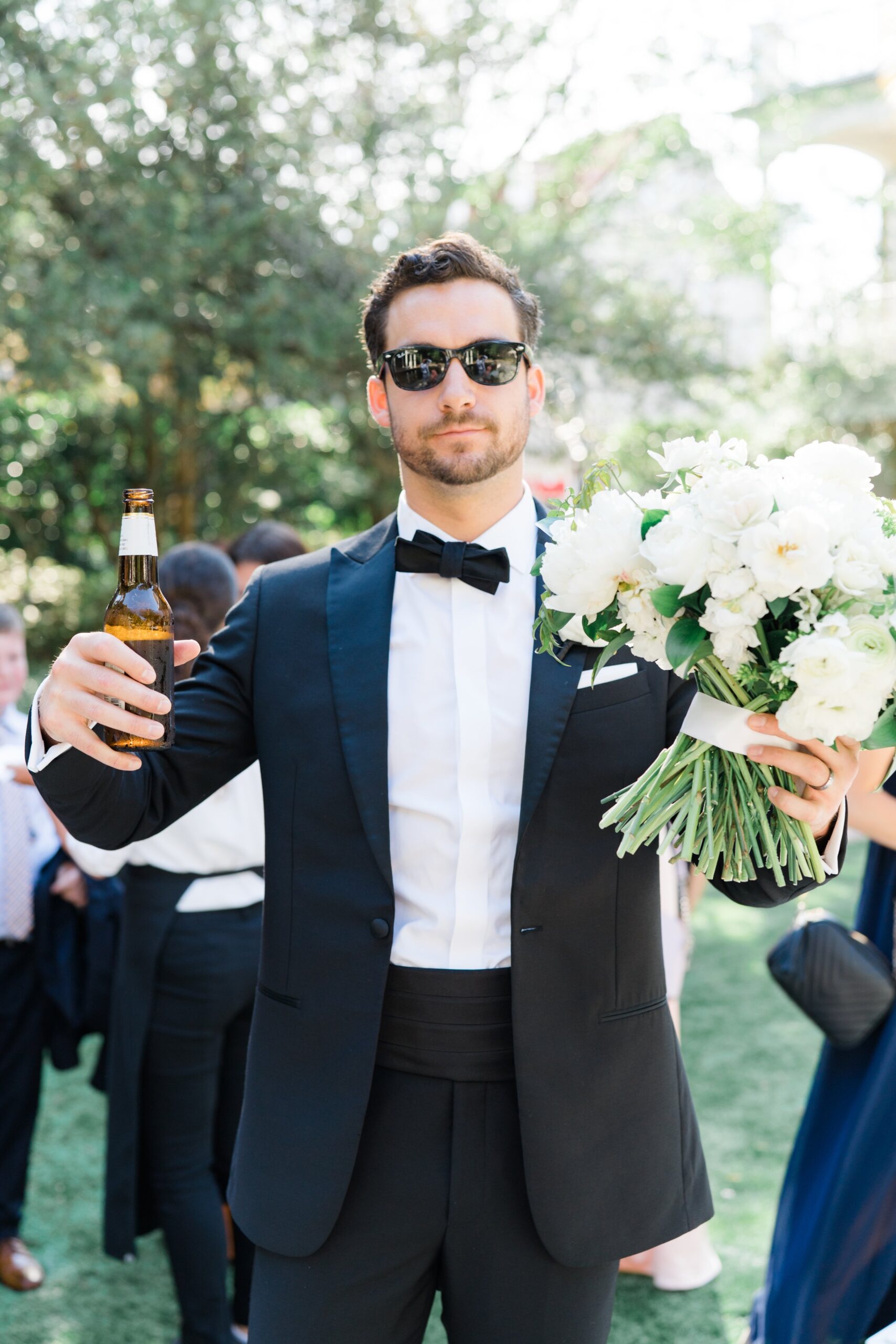 Tuxedo groom holding beer and bridal flowers.