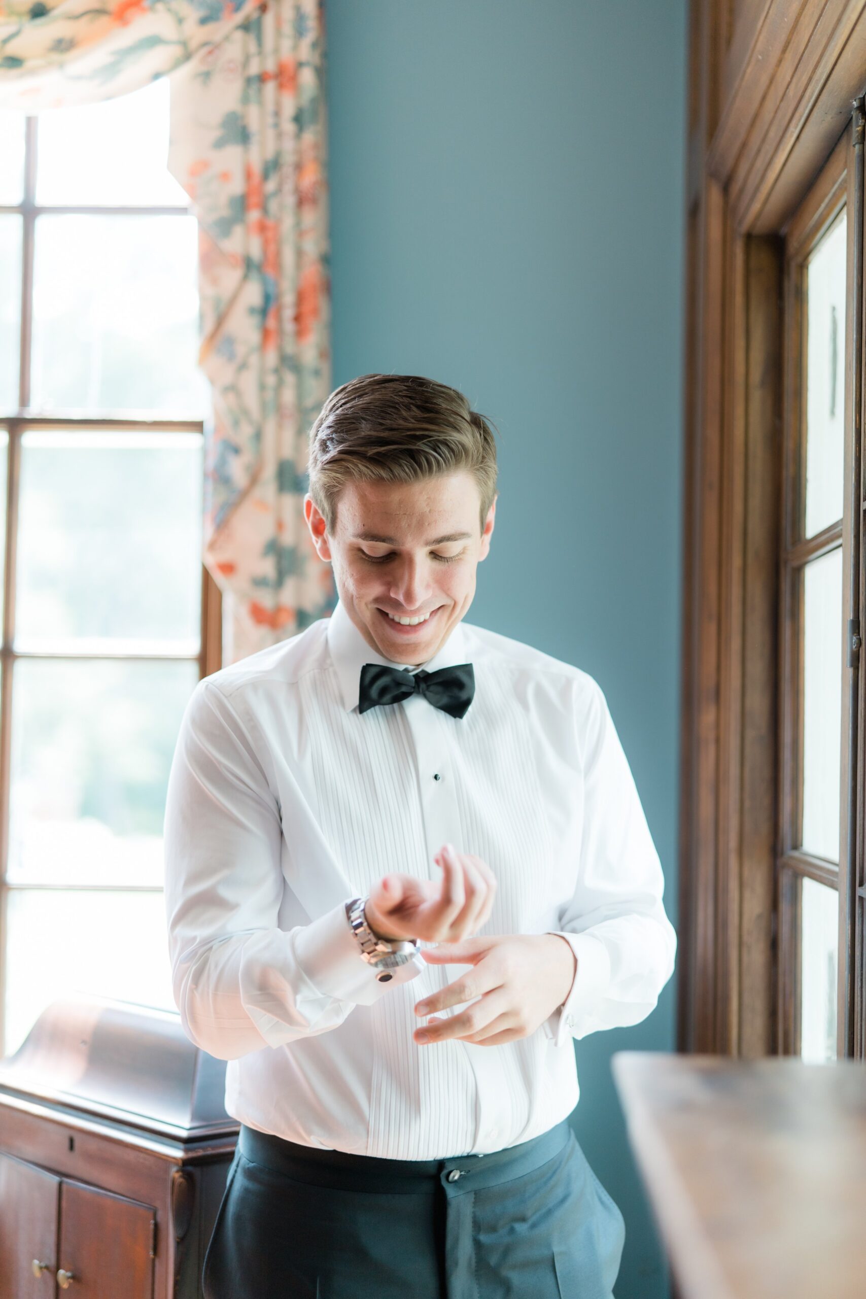 groom in black bow tie smiles and looks down while adjusting cufflinks
