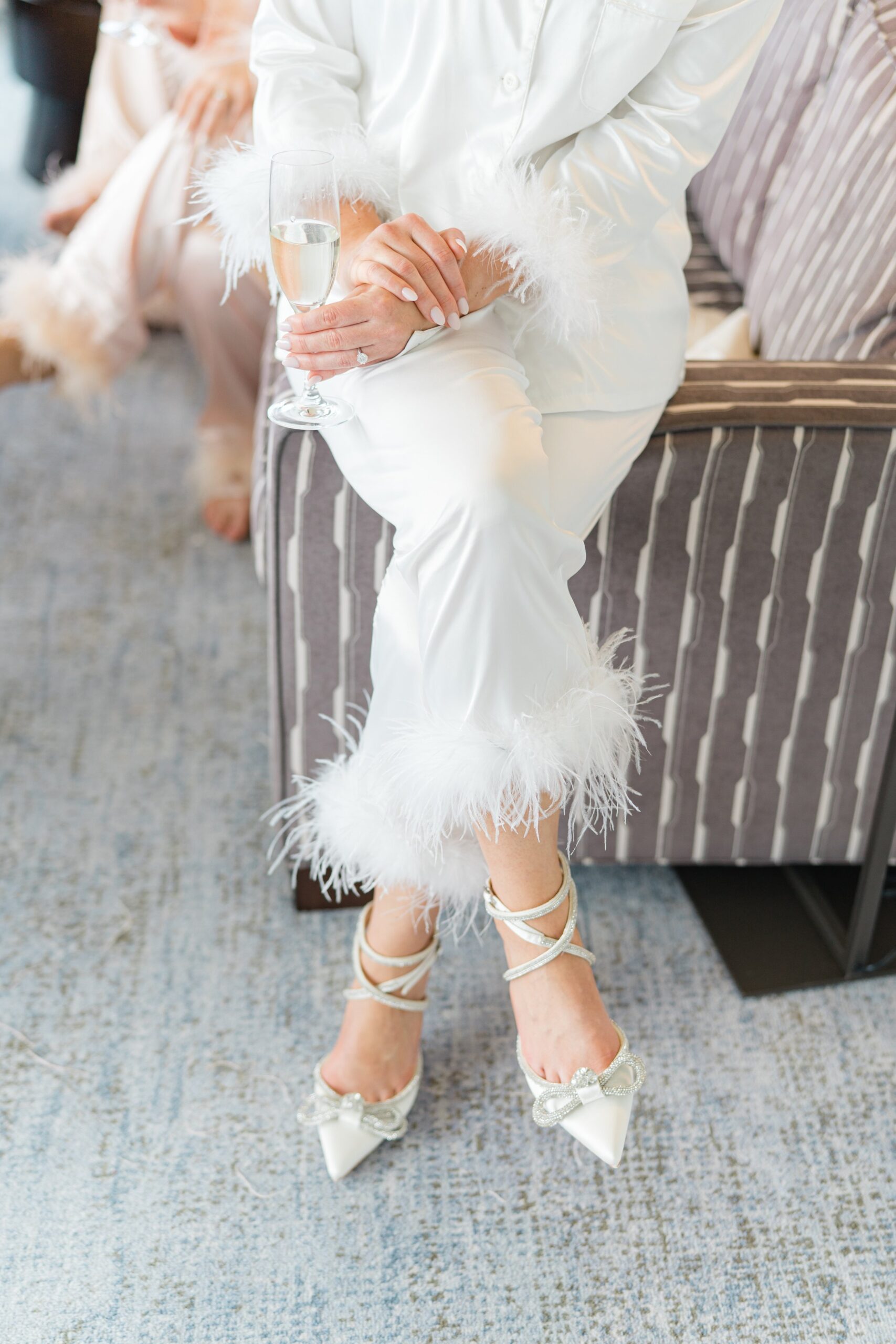 Bride in ankle strapped heels and feather accented white silk pajamas sitting on the arm of a couch and holding champagne.