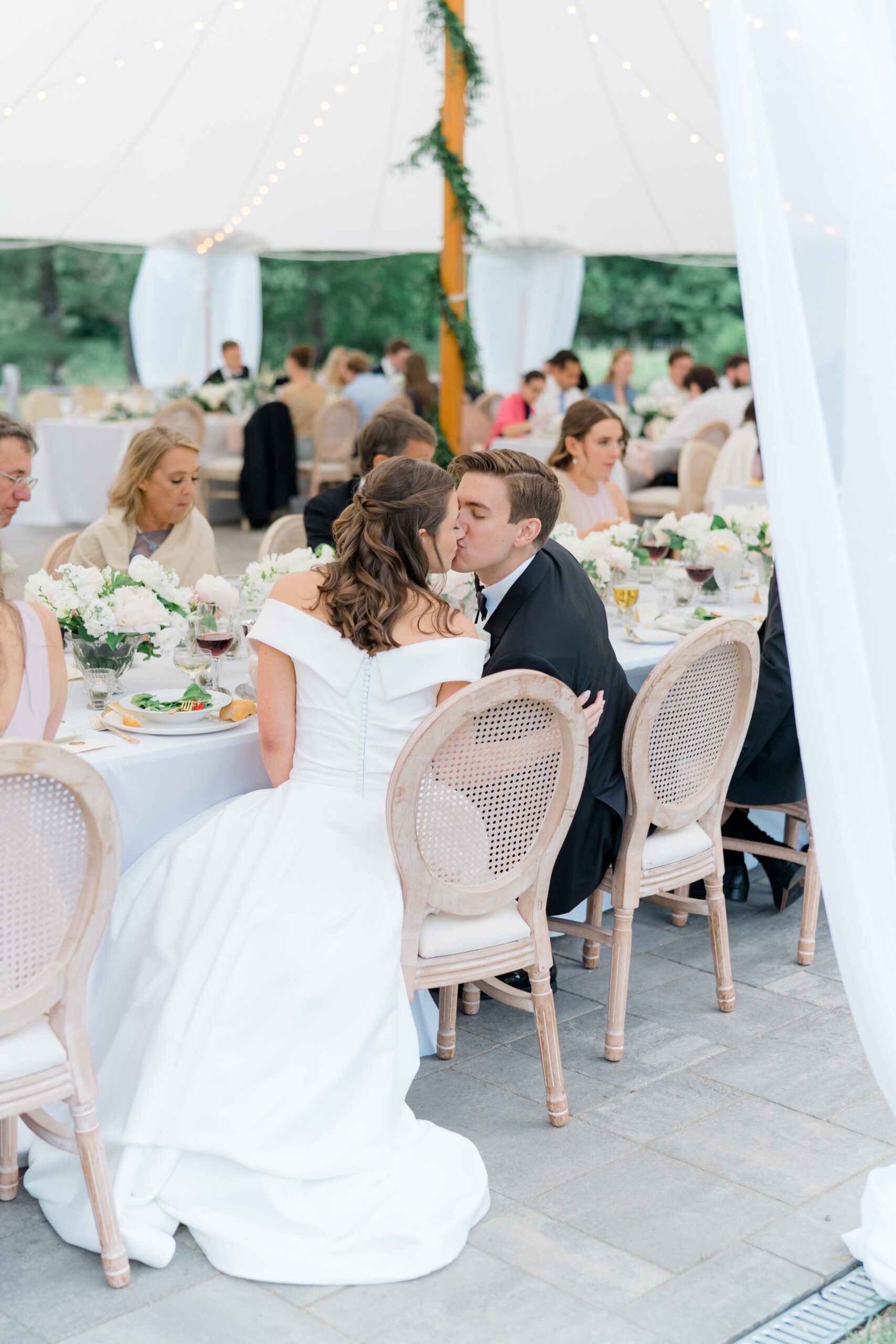 Bride and groom kiss at head table under tent with string lights and lush greenery covered tent pole.
