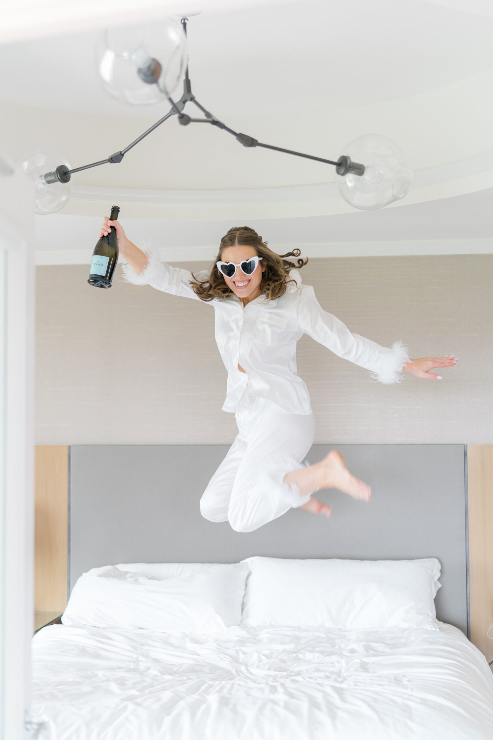 Bride jumps on the bed with prosecco bottle in one hand while wearing white heart shaped sunglasses. Boston weddings.