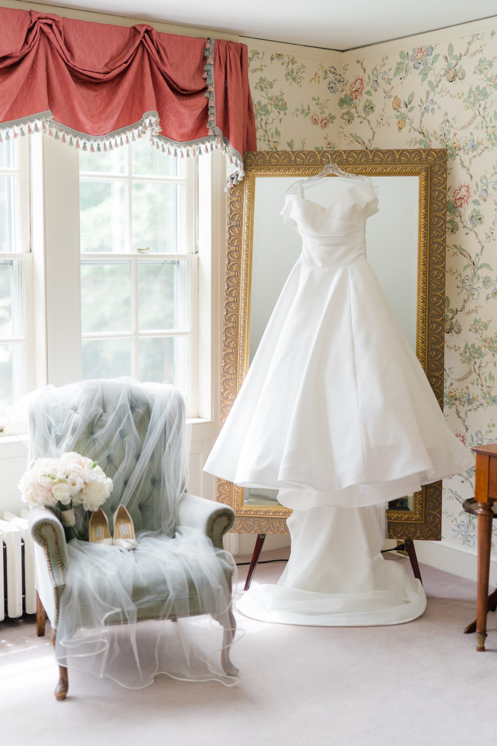 Wedding dress hanging on golden framed mirror with heels, flowers, and veil. Bradley Estate Bridal suit. Classic wedding venue in Boston