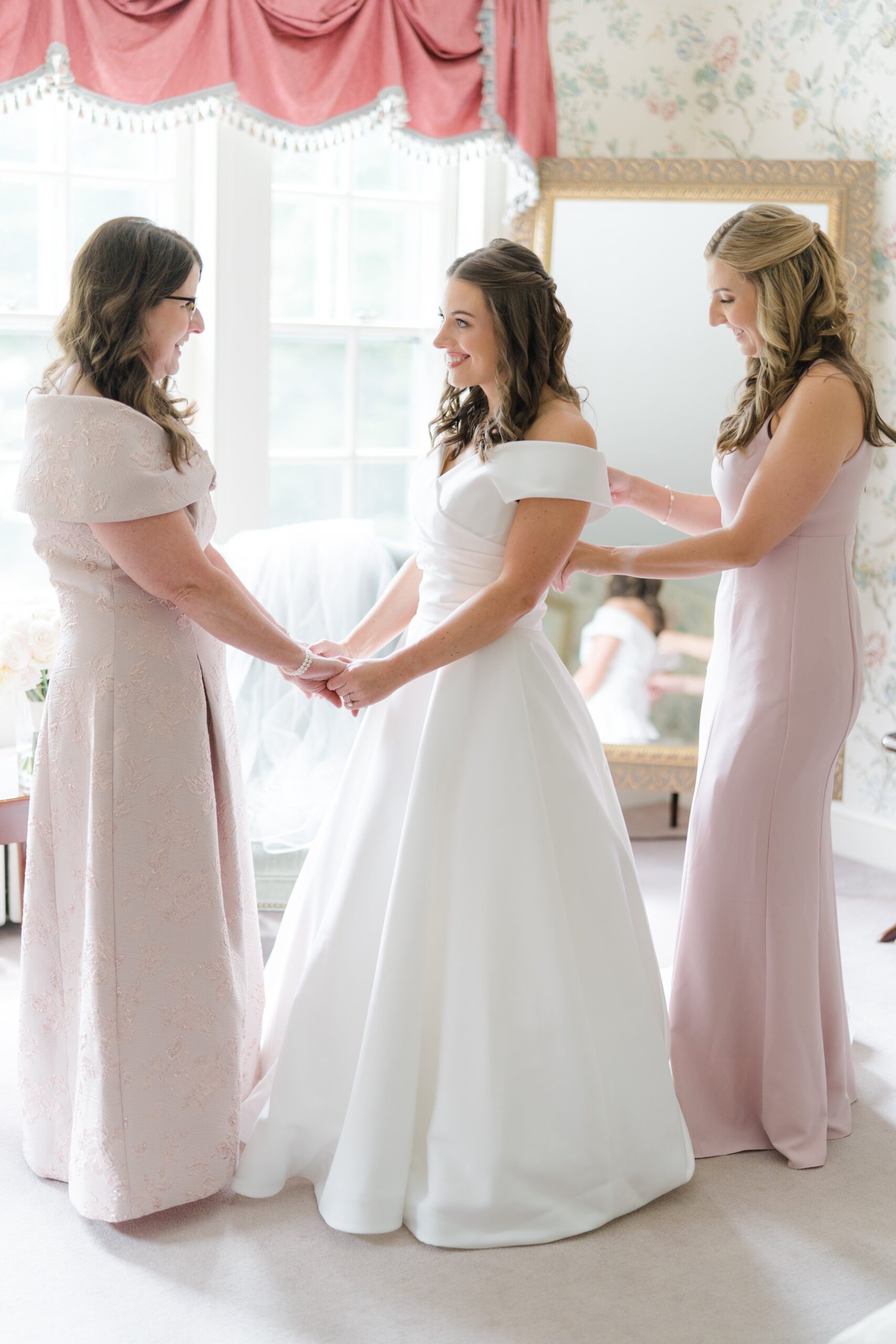 Bride and her mom share a moment while maid of honor finishes buttoning wedding dress.