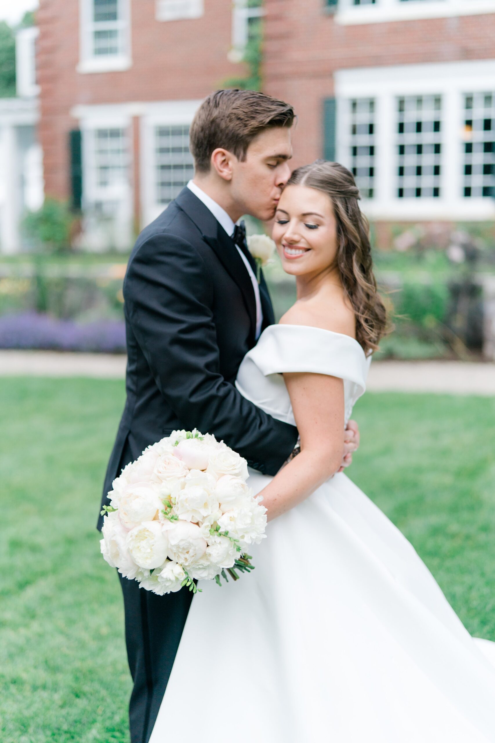 Groom kisses the temple of bride holding classic white flowers
