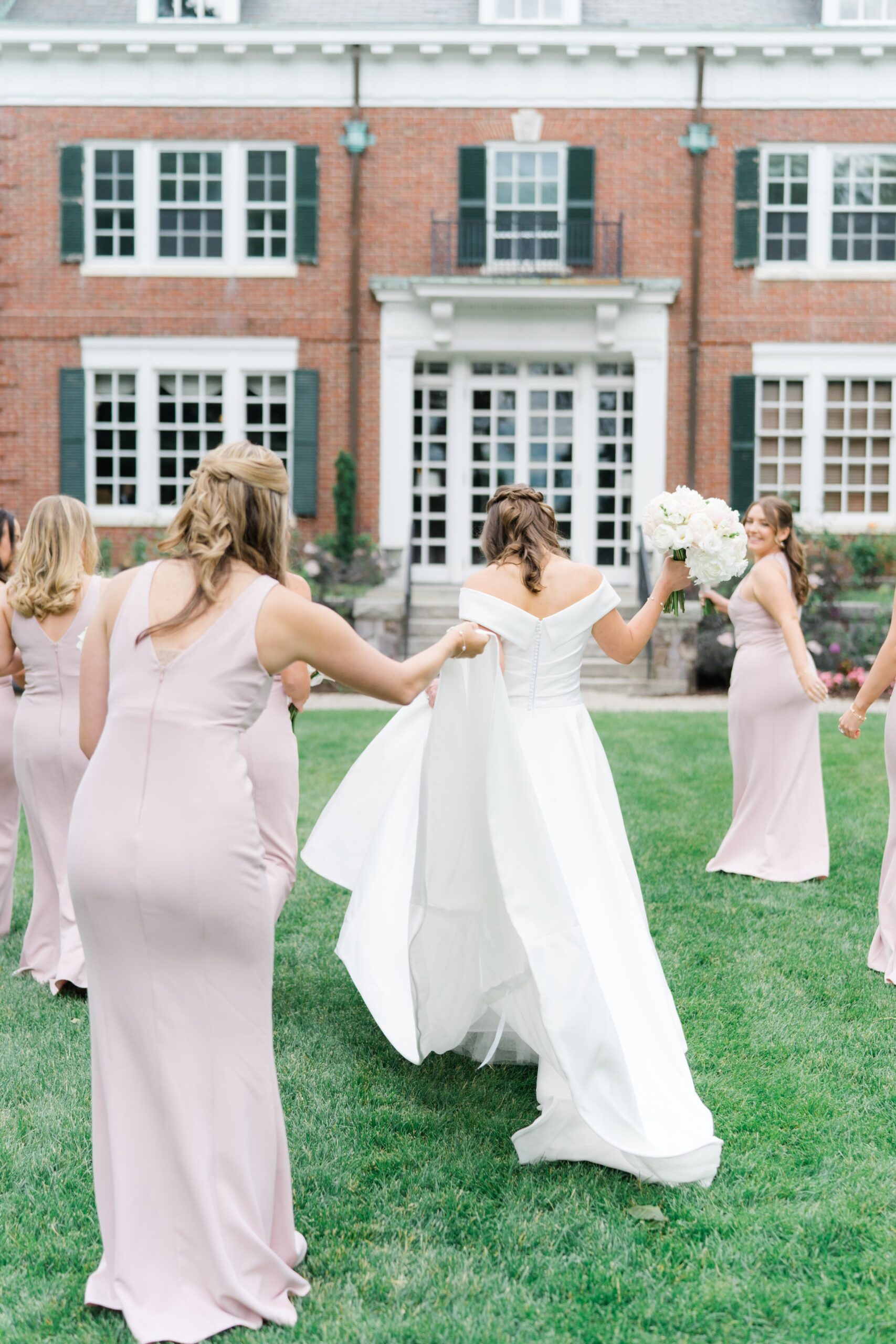 Maid of honor helps bride with dress while walking on the lawn.