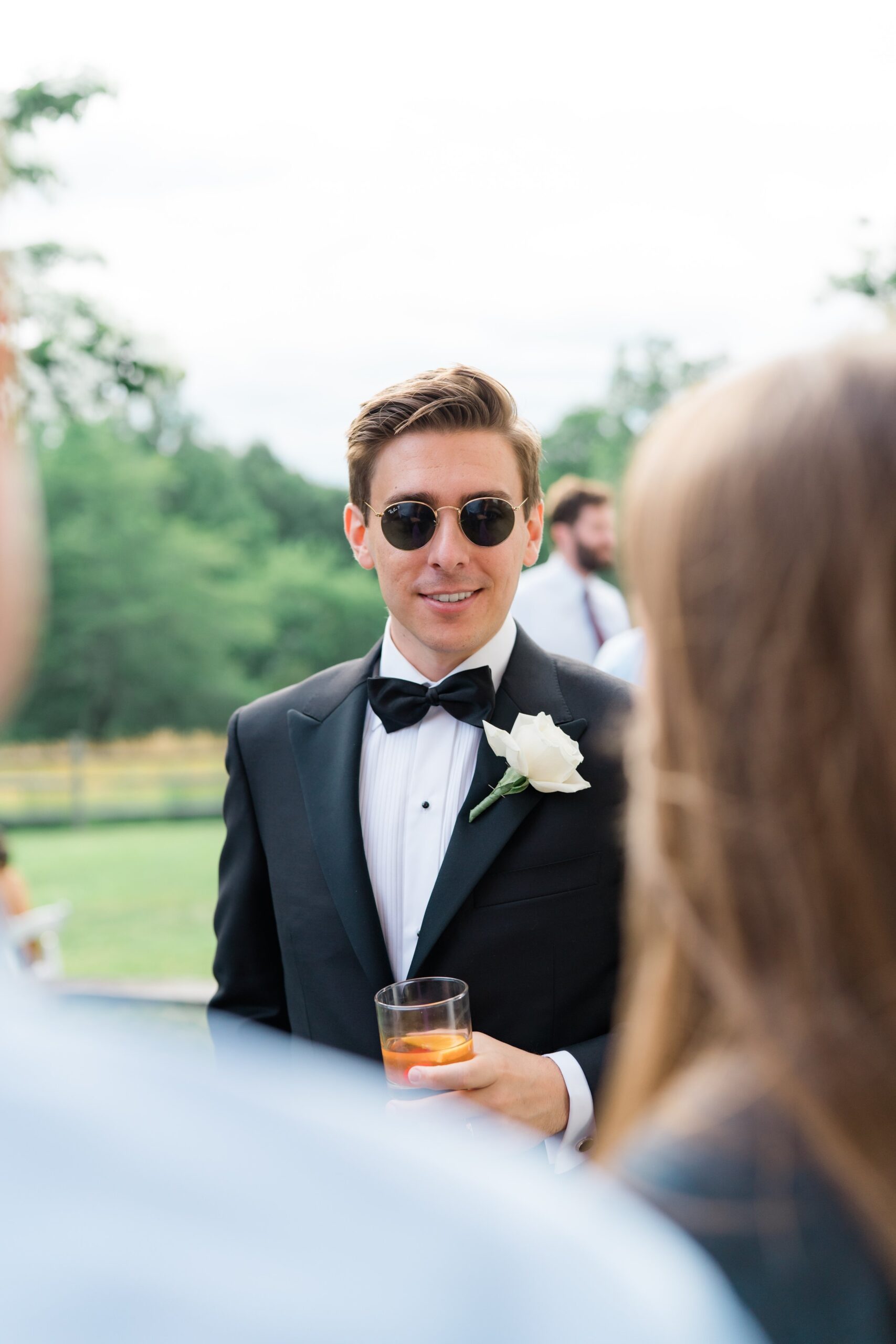 candid moment of the groom at cocktail hour wearing circular gold rimmed ray bans and holding an old fashioned. Black tie groom with large white flower boutonniere. New England Destination Wedding Photographer.