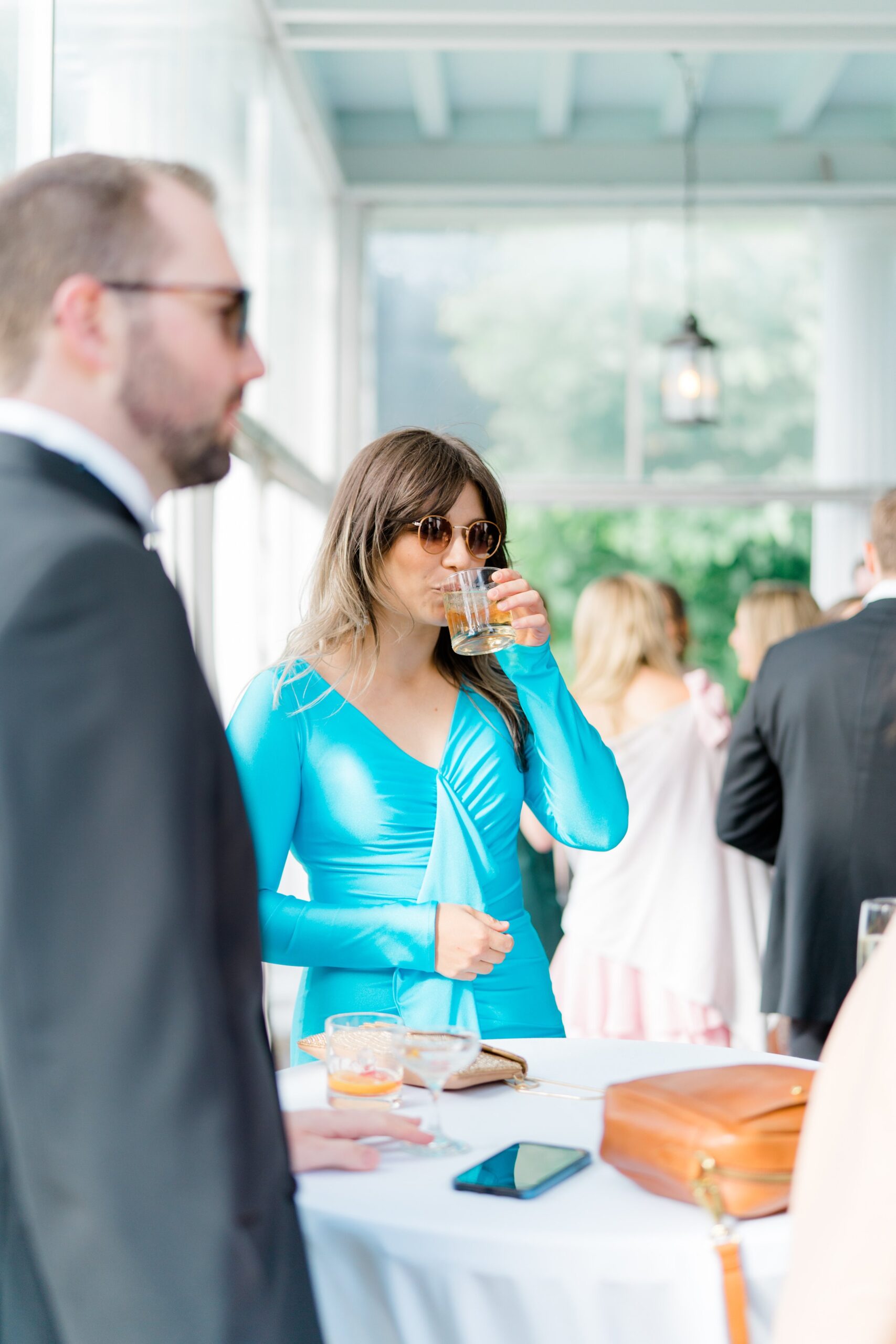 Wedding guest sips drink while wearing bright blue long sleeved dress at Bradley Estate Summer Wedding