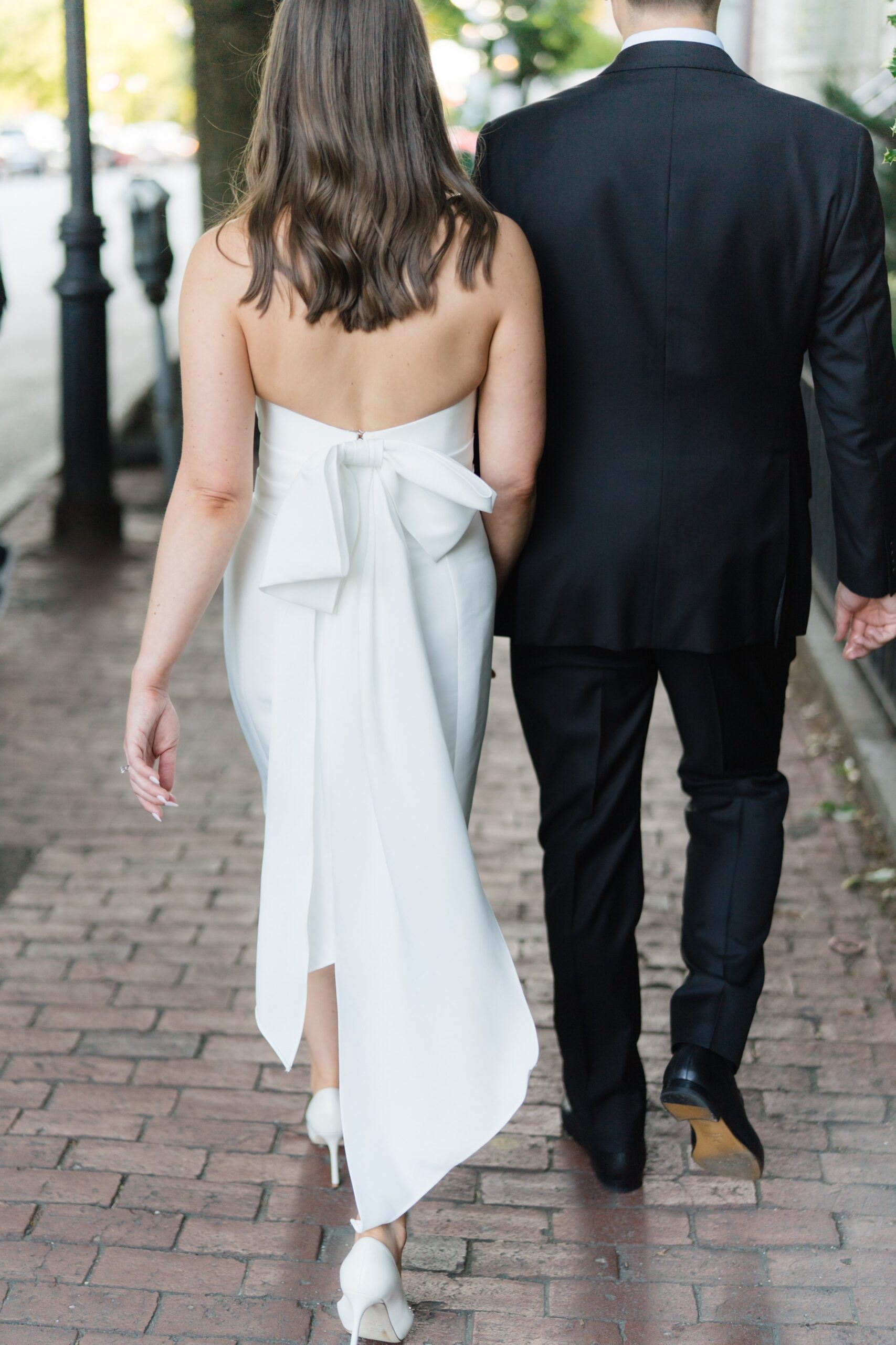 Rehearsal dinner dress for bride with big bow flowing in the wind. Bride and groom holding hands walking down the street.