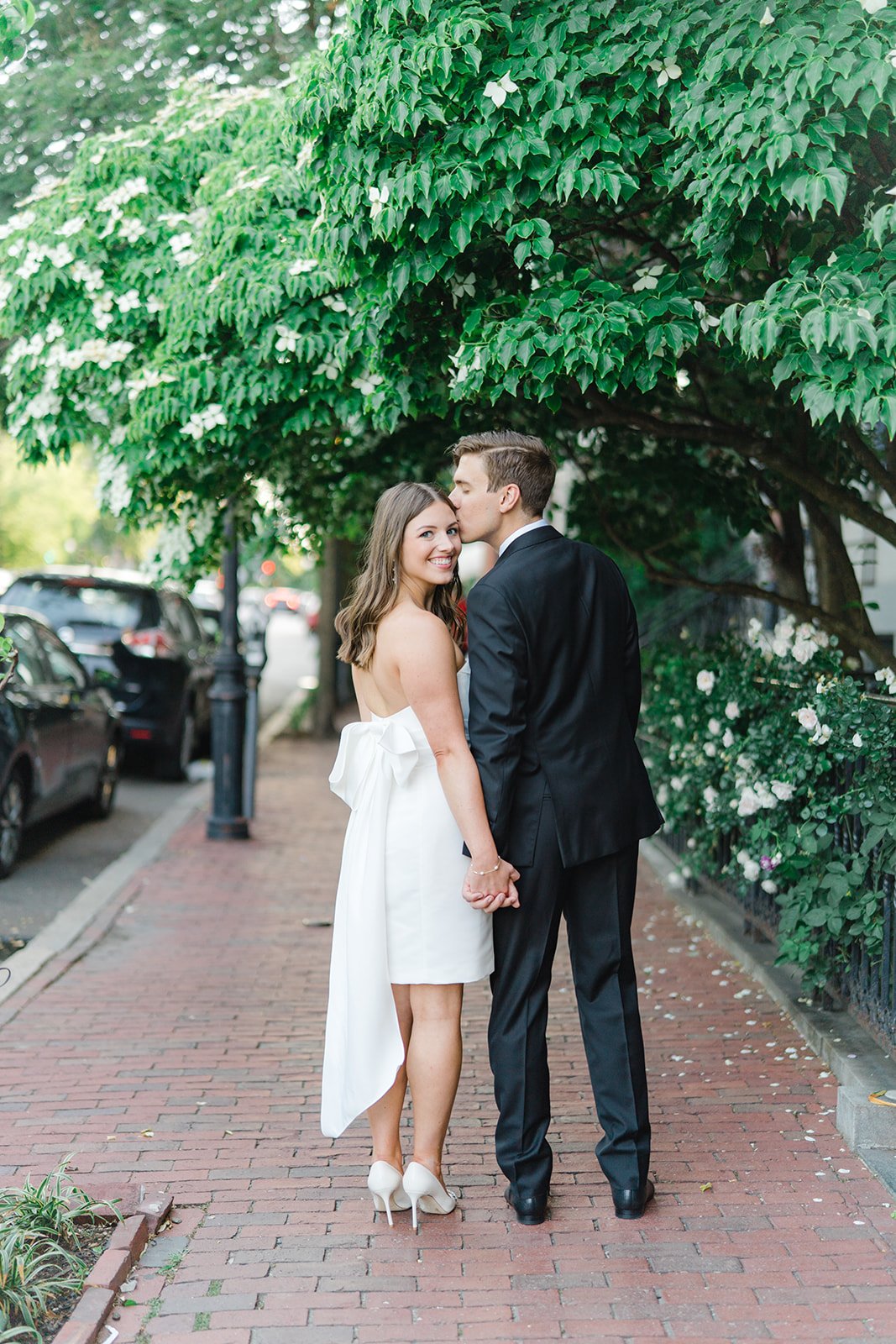 Cute strapless rehearsal dinner dress with large bow with long tail. Groom kisses bride to be on cute sidewalk with spring tree in downtown boston.