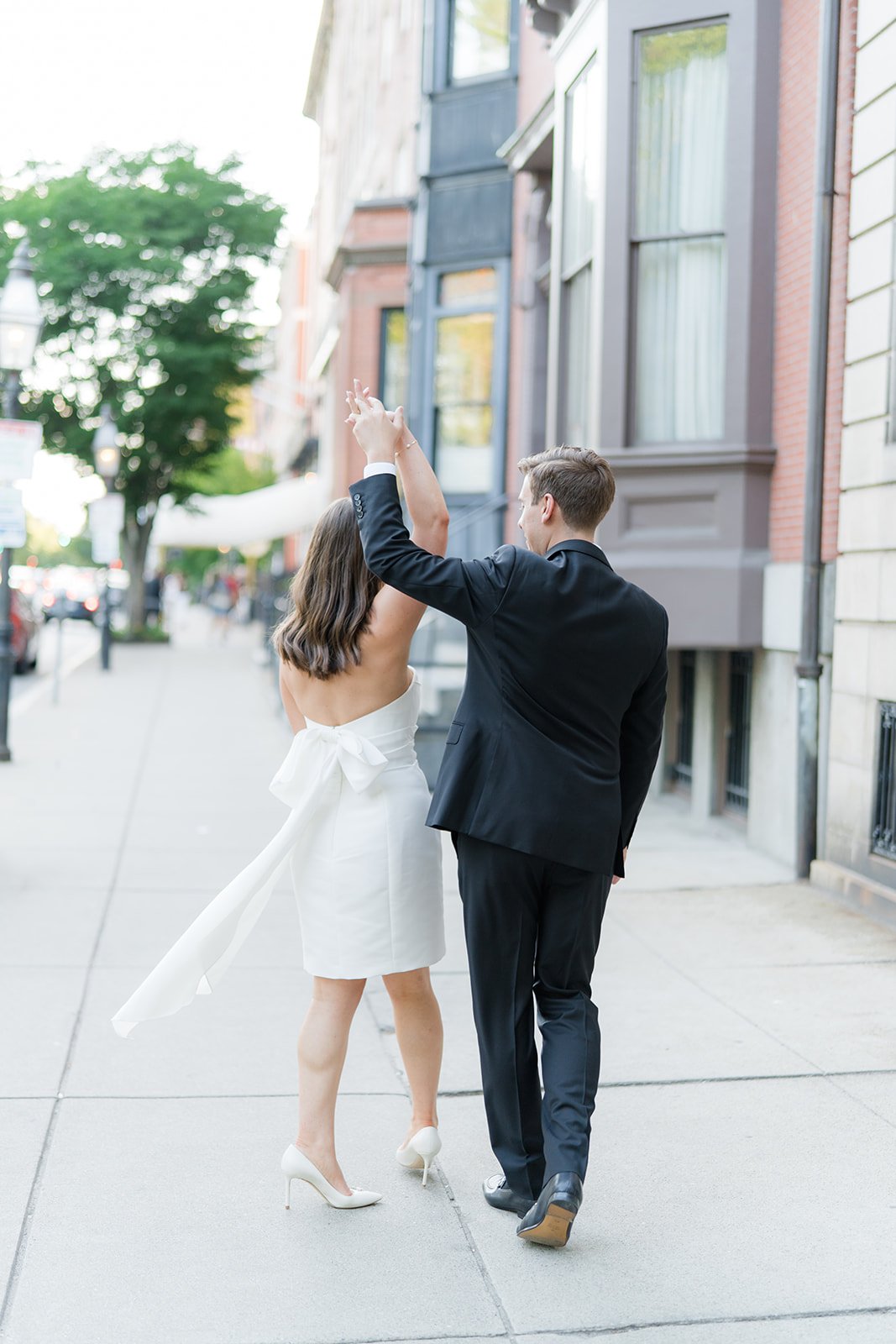Walking on the sidewalk in Beacon Hill. Groom twirls his bride to be with her bow flowing in the wind. Summer wedding in boston.