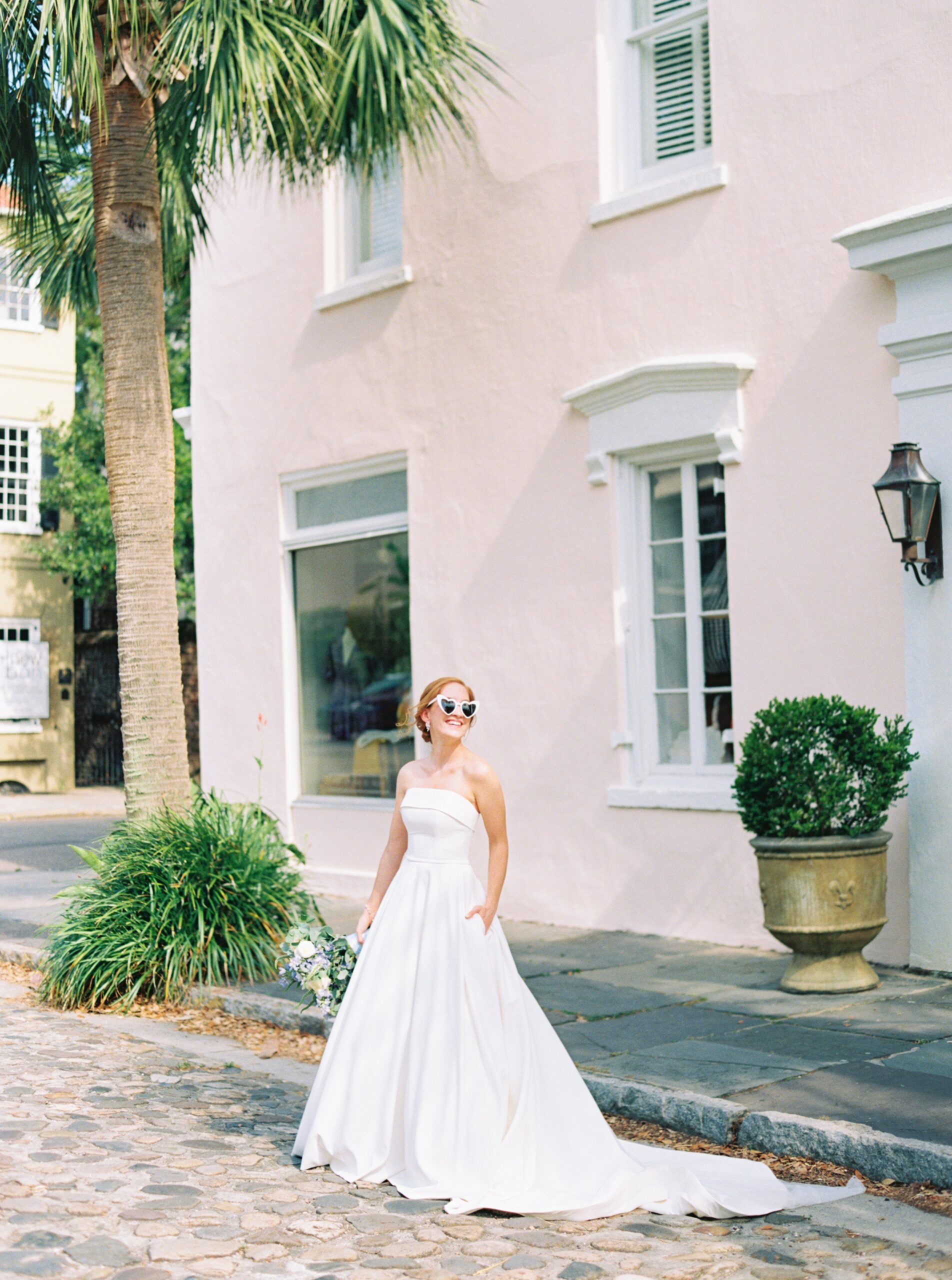 bride in strapless dress and heart shaped sunglasses standing on cobblestone street with pink wall and palm tree. top charleston wedding photographer. wedding dress with pockets.