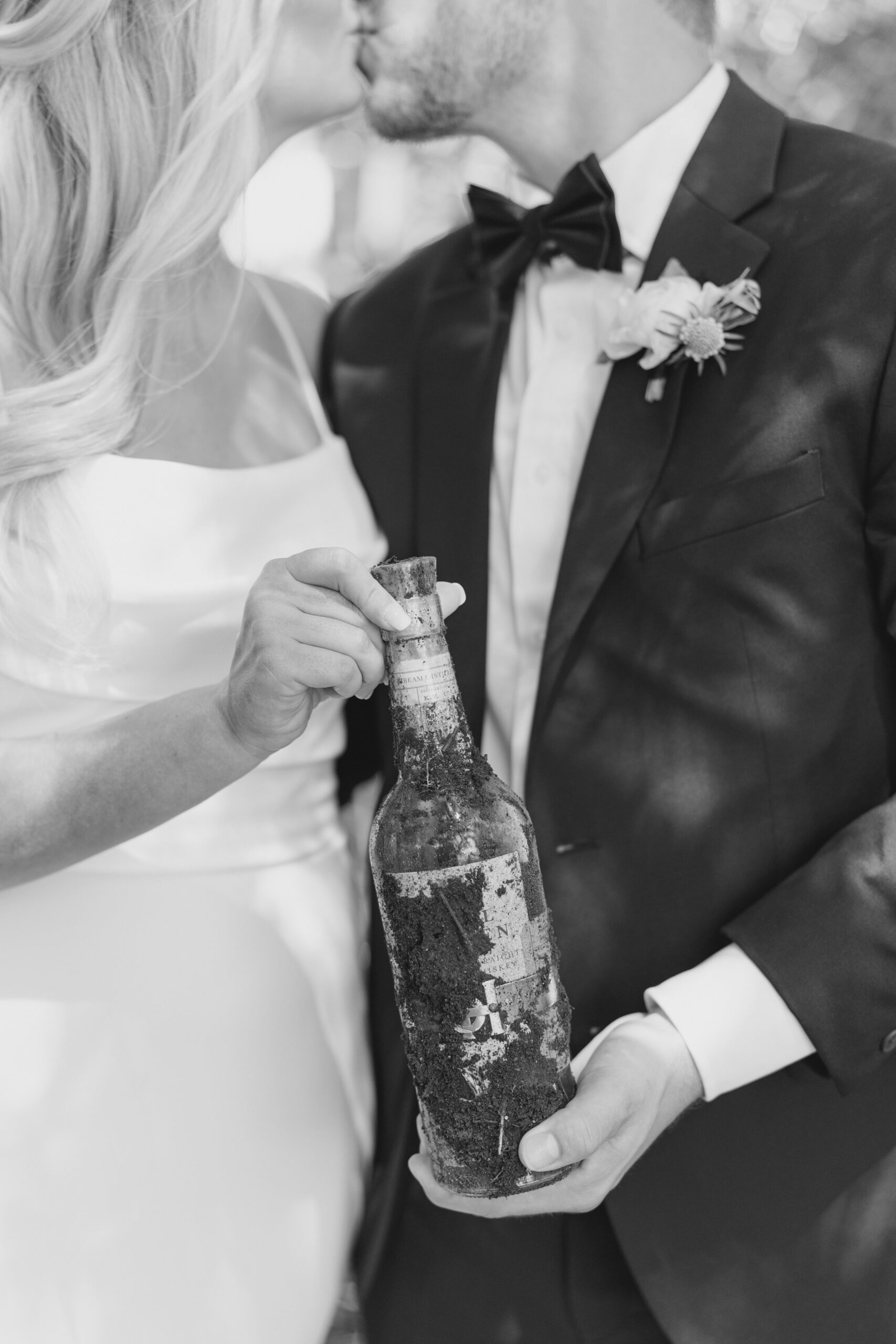 black and white wedding photo. bride and groom kiss while holding dirt covered bottle of bourbon.
