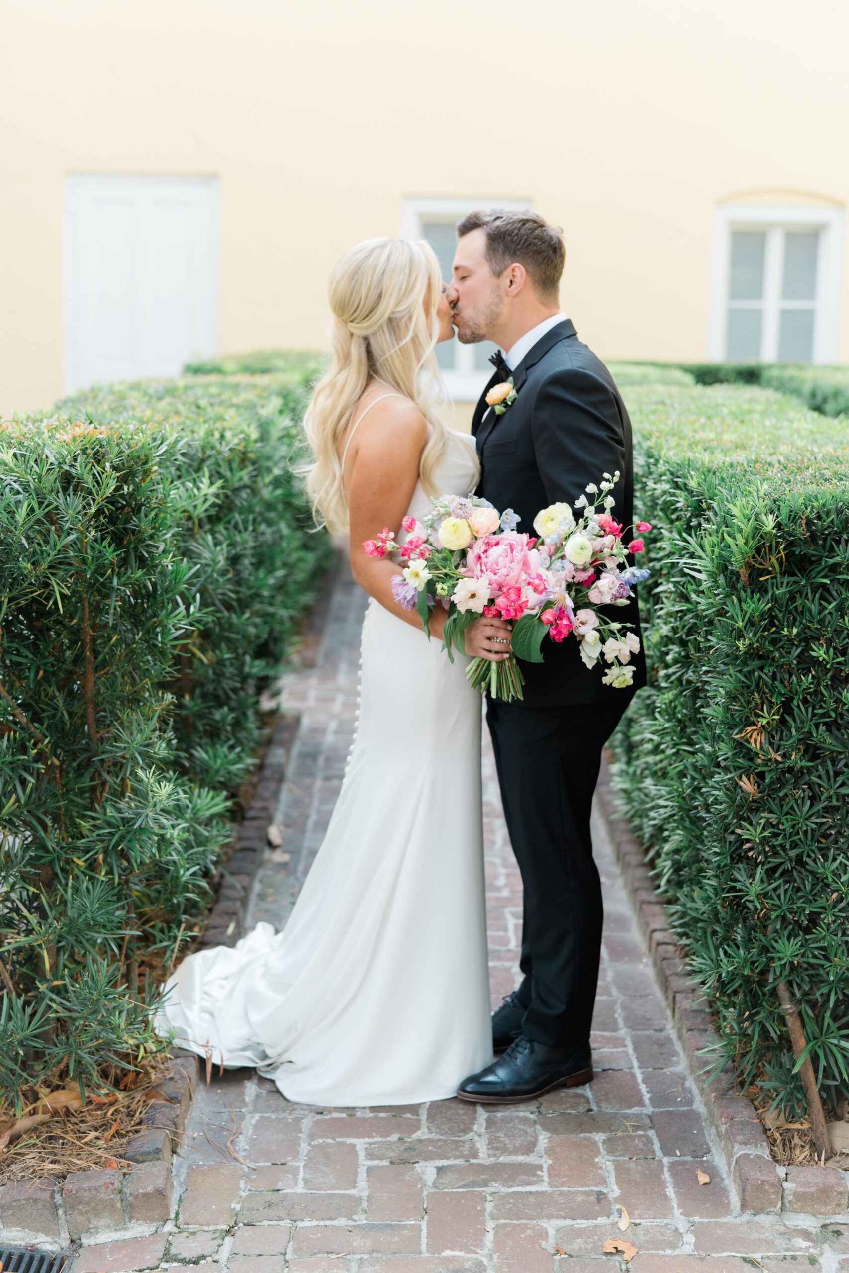 bride and groom kiss between green bushes with yellow wall