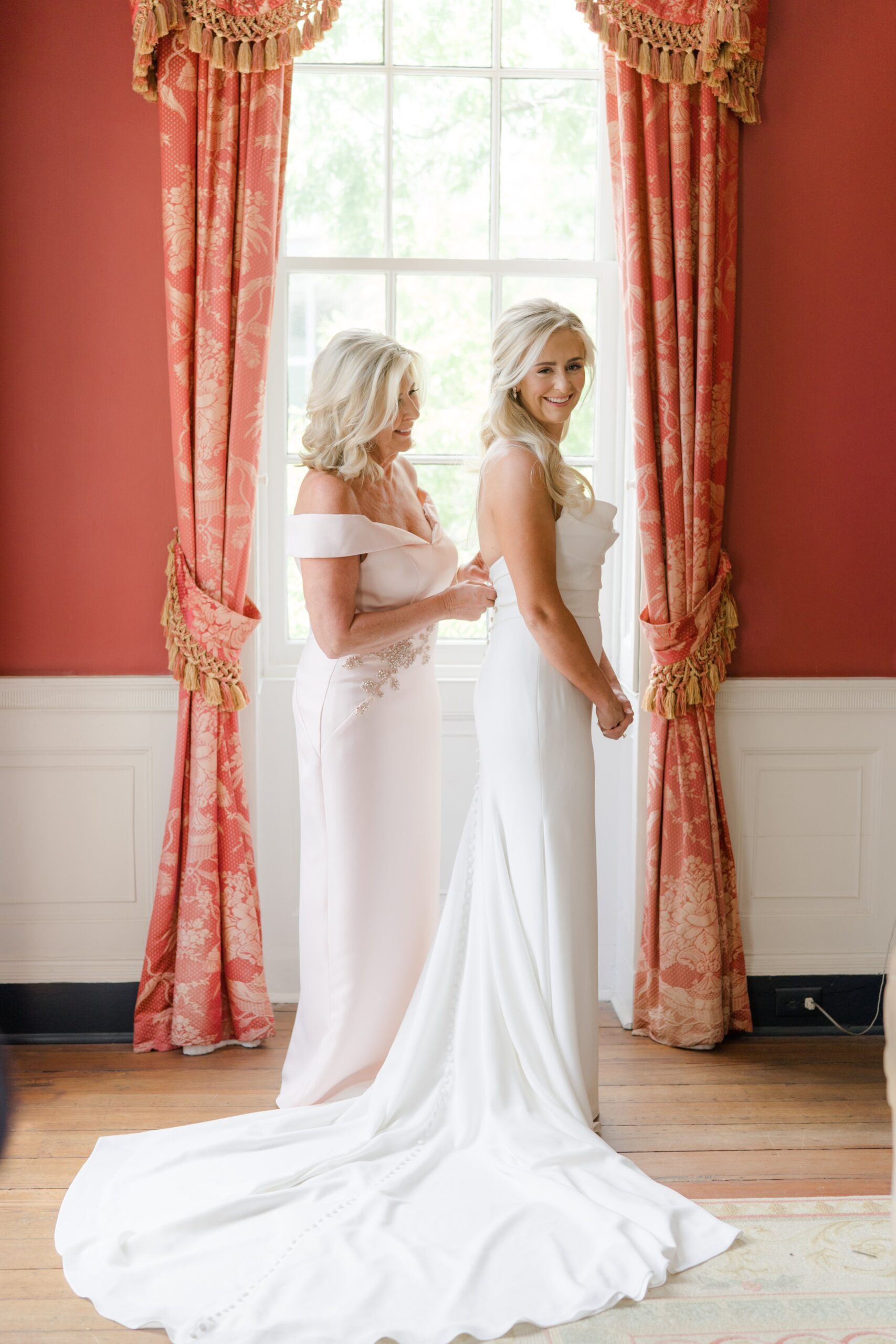 mom helps blonde bride get into wedding dress in red room with gold lined drapes at william aiken house.