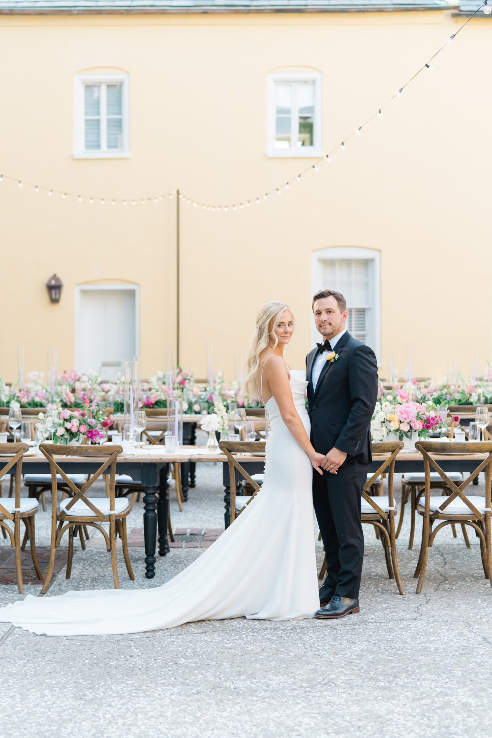 bride and groom softly holding hands with yellow wall, string lights and al fresco dining reception set up