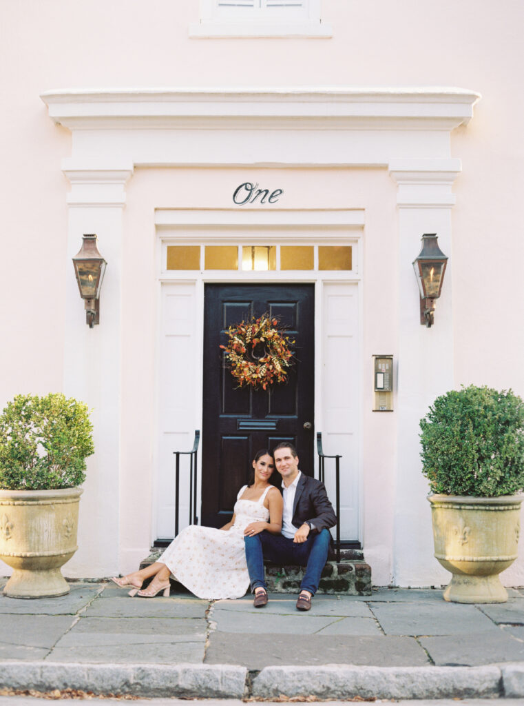 couple sitting on steps of pink building with gas lanterns. Downtown Charleston engagement. Greenery bushes. 