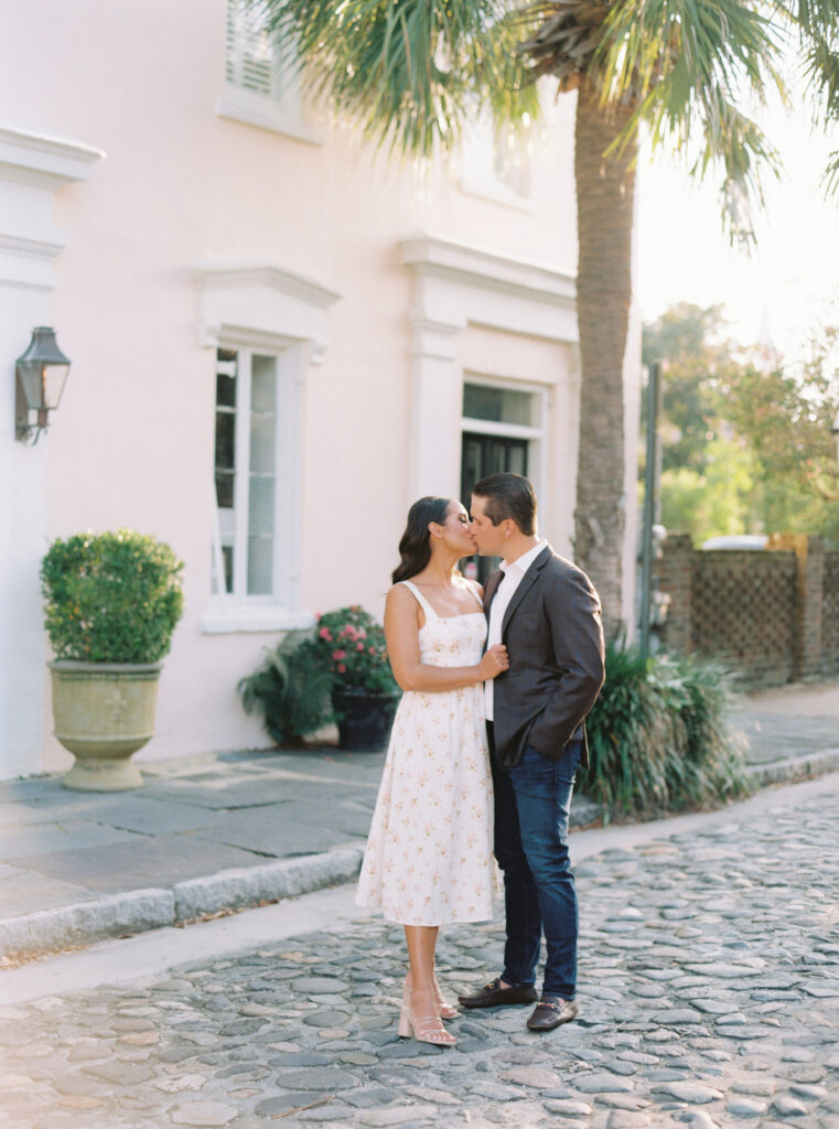 Couple kissing on cobblestone street with palm tree and pink wall. 