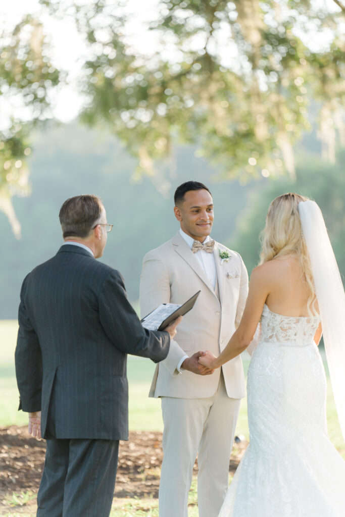 Groom holds hands and looks at bride during wedding ceremony. 