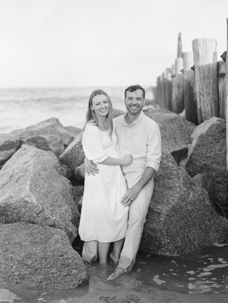 Black and white photo of a couple smiling and sitting on rocks at the beach. Photographer weddings Charleston. 