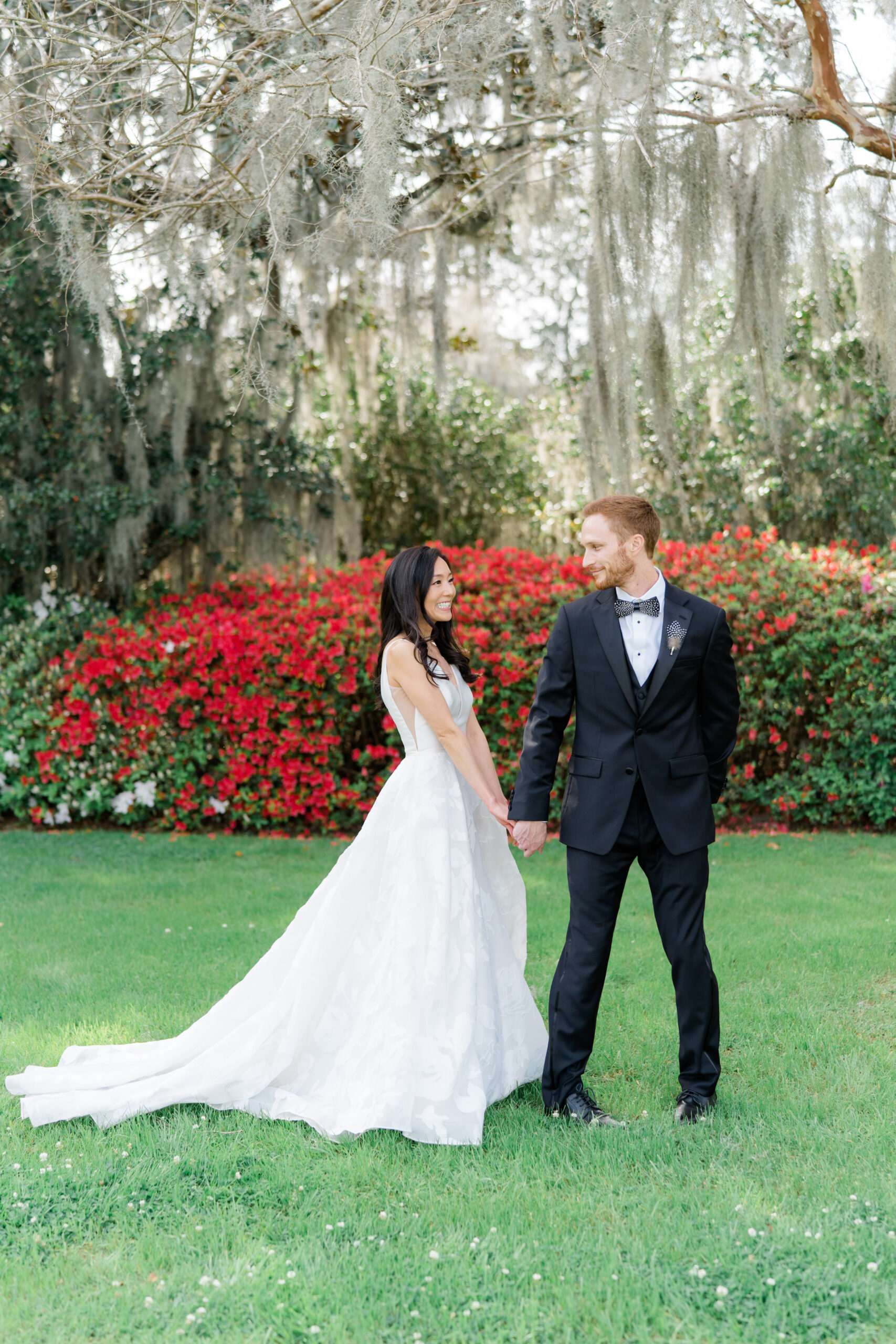 Groom smiles as he sees the bride during first look. Red azalea blooms and spanish moss. Middleton Place gardens. 