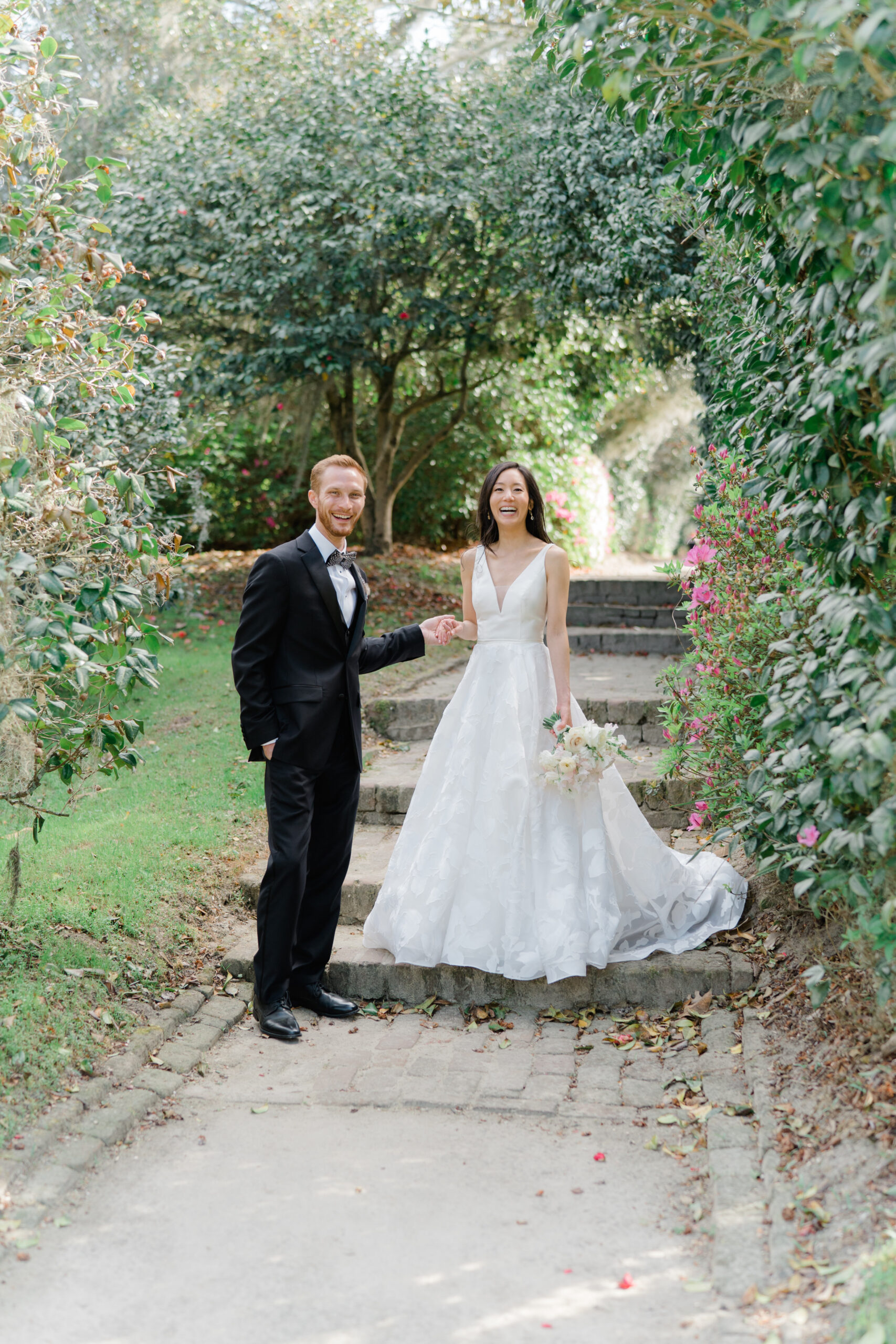 Groom leads the bride down the steps in Middleton Place gardens. Lush greenery and flower blooms. charleston photographer wedding.