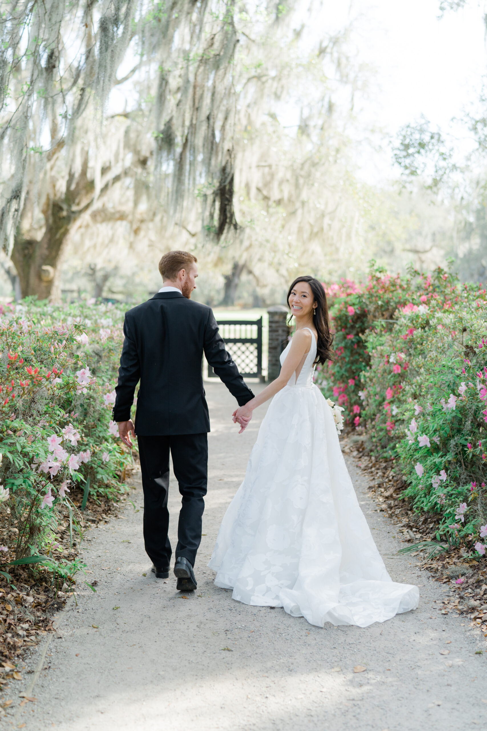 Bride looks back at camera while walking with groom on a path at Middleton Place with spanish moss blowing in the wind. 