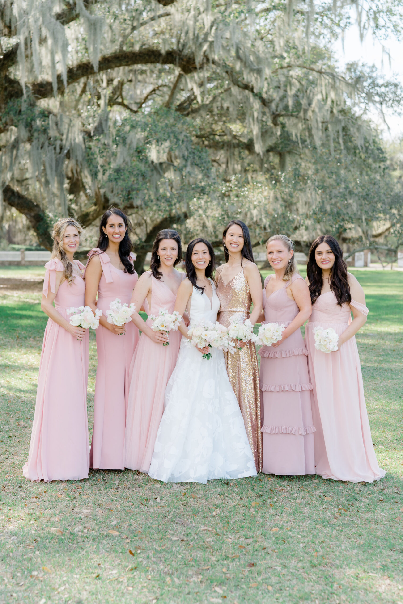 Bride and bridesmaids group photo. Bridesmaids wearing pink and the maid of honor wearing a shimmering gold sequins dress. Live oak trees and spanish moss. Middleton Place wedding.