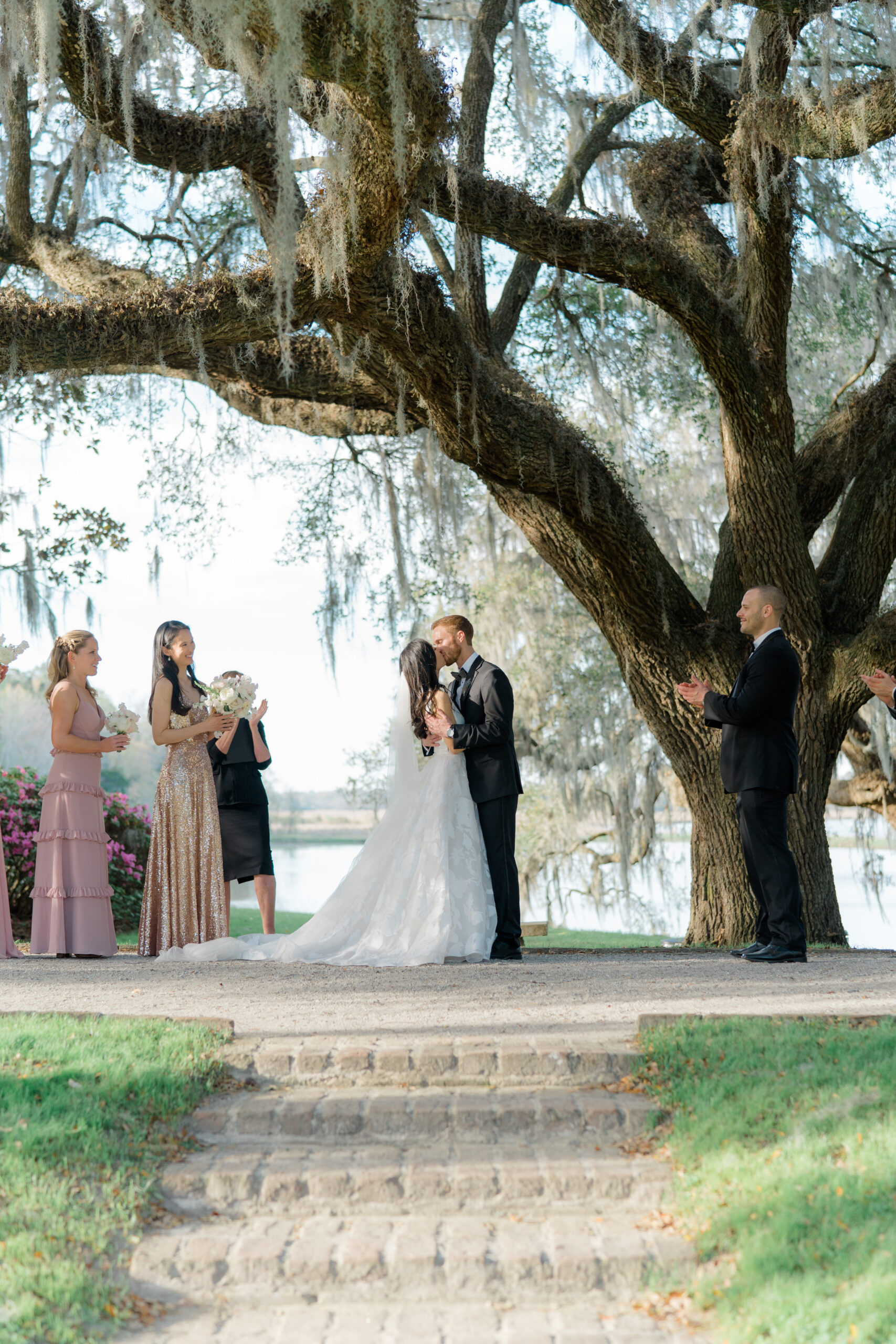 First kiss under the tree with spanish moss during wedding ceremony in the octogonal garden at Middleton Place. Bridal party clapping. Maid of honor in shimmering gold dress. 