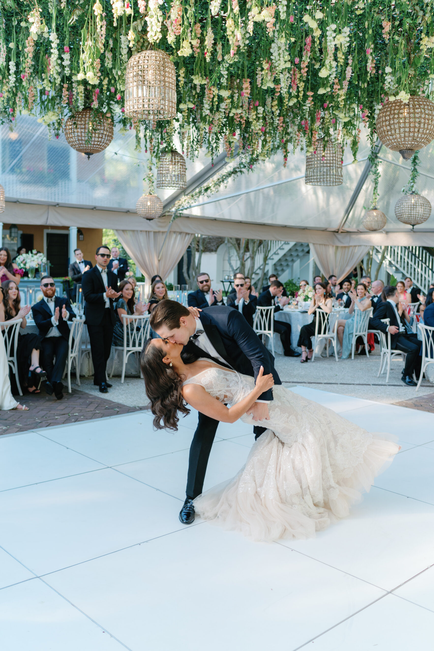 Wedding first dance dip kiss on all-white dance floor with flowers hanging above. 