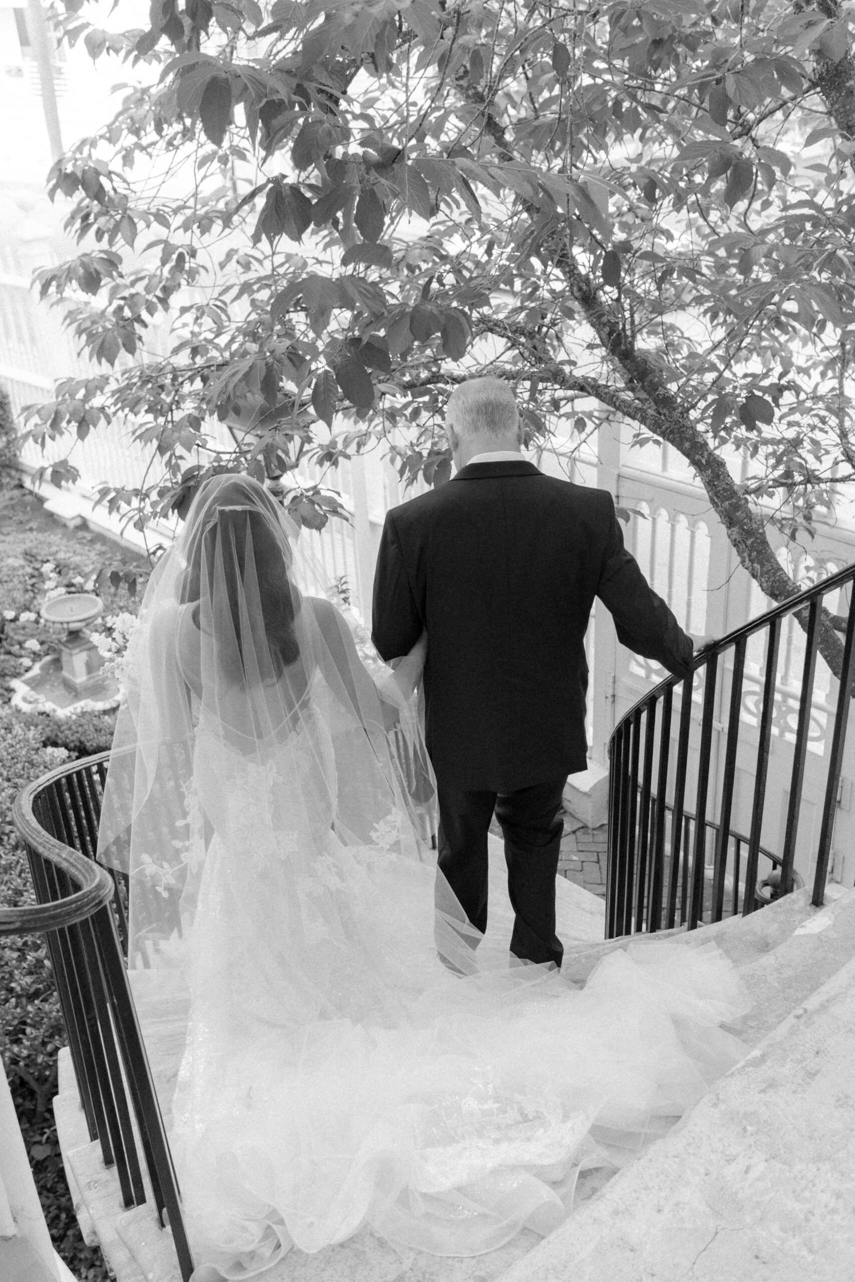 Black and white photo of bride walking down curved marble steps with her dad.