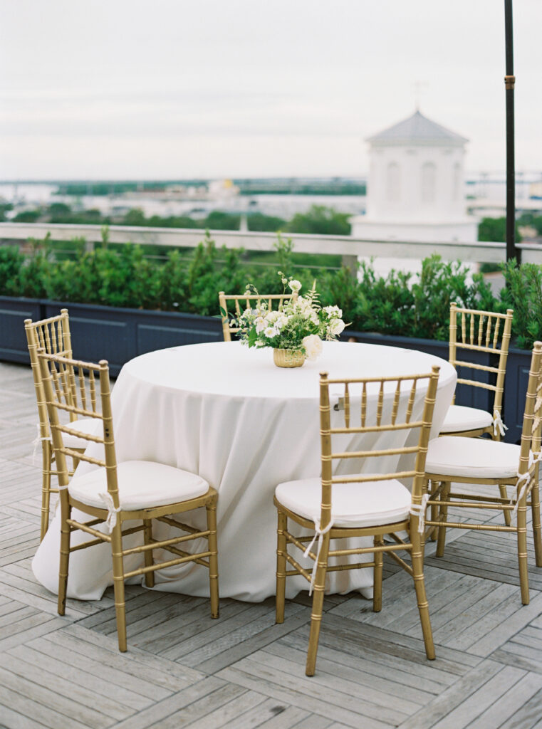 Charleston rooftop welcome party. White table cloth, white and green flowers, gold chairs. 
