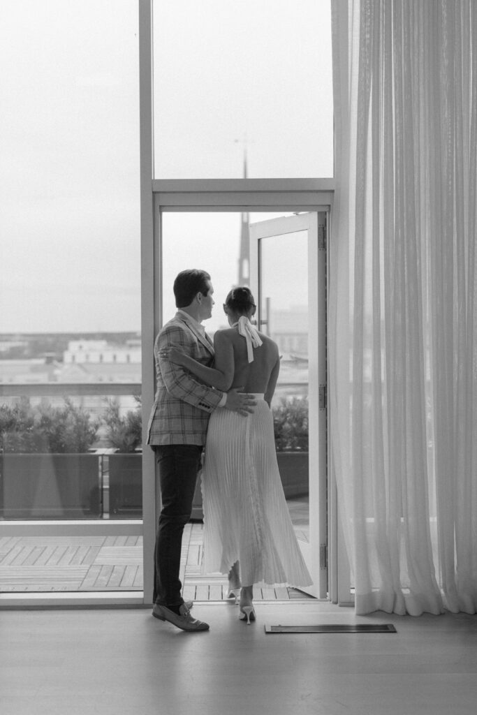 Black and white romantic photo. Groom holding door open for bride with one hand on her back. Charleston church steeple in the background. Rooftop welcome party.