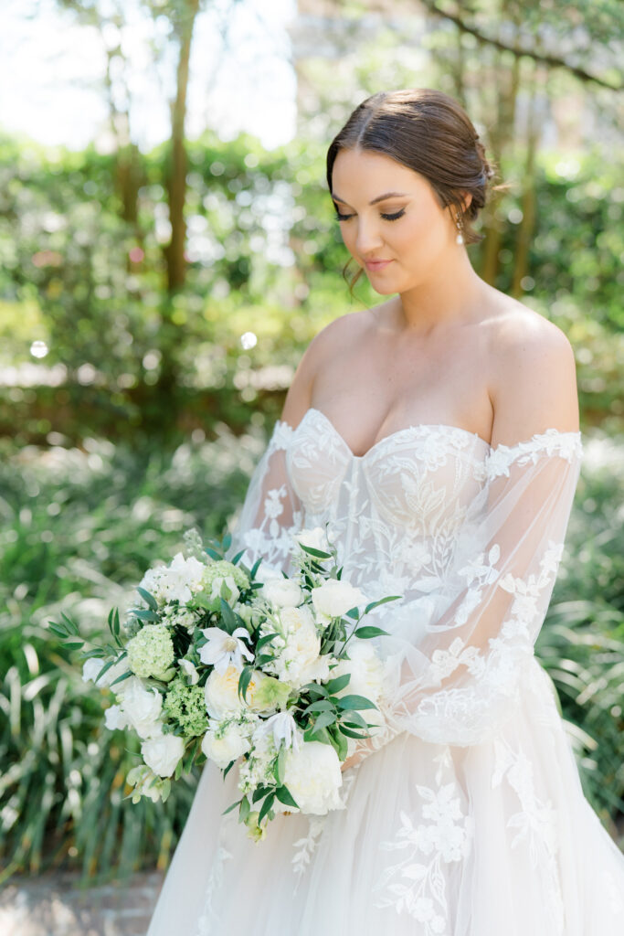 Bride in strapless dress and detachable sleeves looking down at white and green bouquet. 