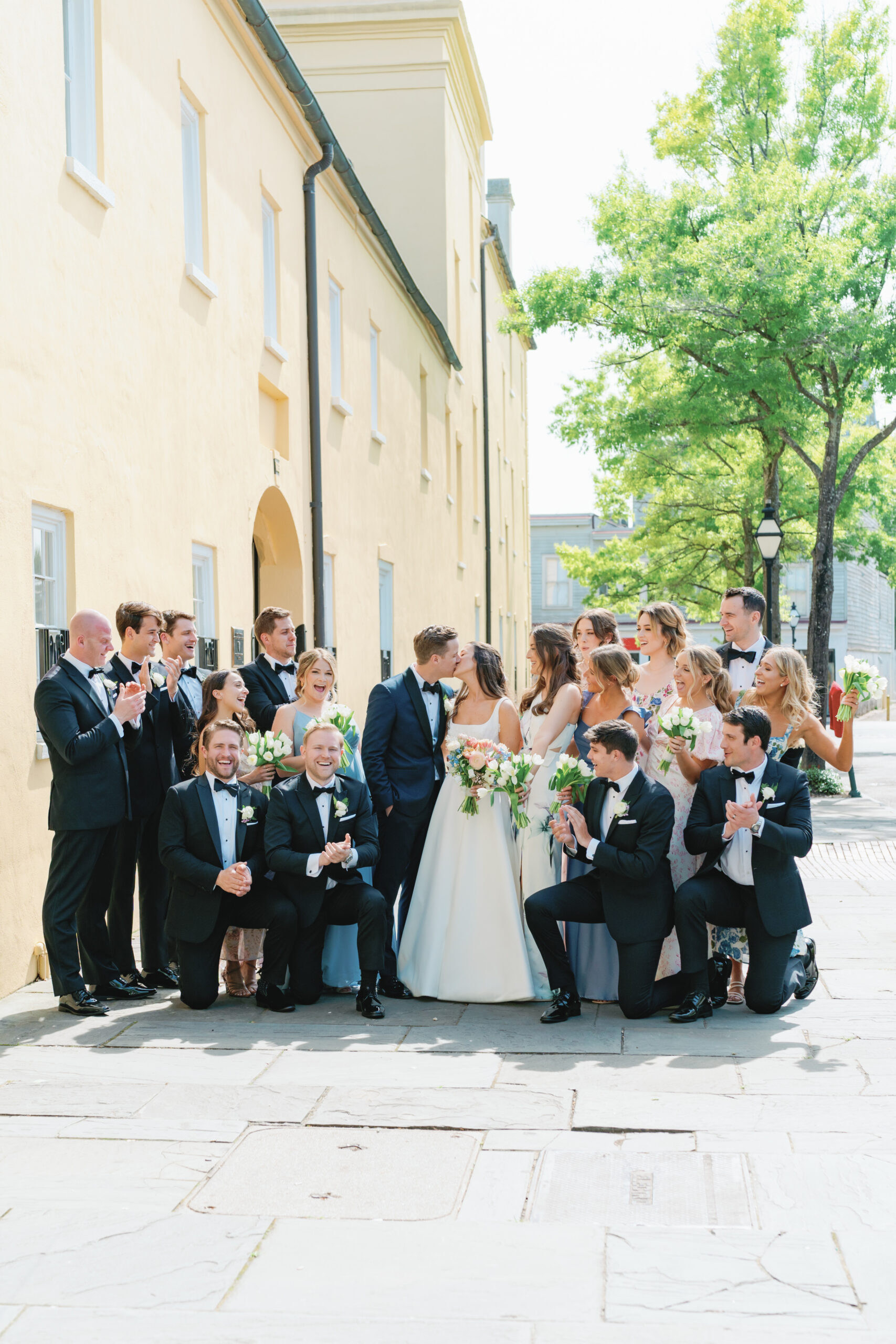 Full bridal party on the sidewalk in downtown Charleston. Big yellow wall and green trees. spring destination wedding. 