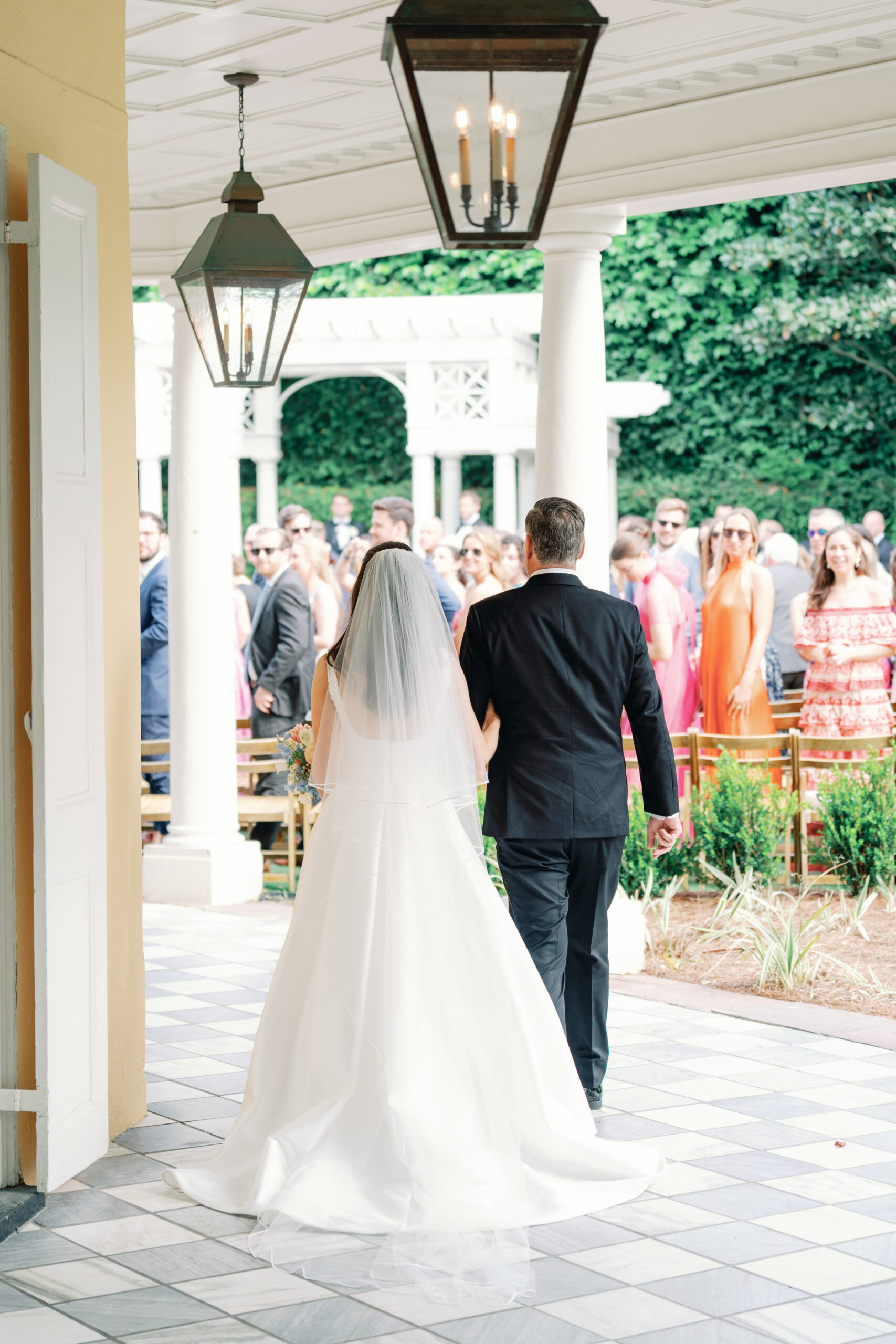 Bride walking to wedding ceremony escorted by her dad. Checkerboard marble floor. Garden ceremony in Charleston, SC.