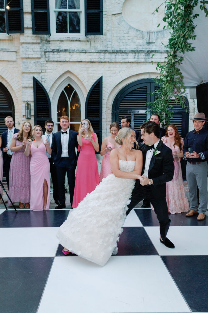 Bride and groom lean to the side during first dance with bridal party smiling and laughing in the background. 