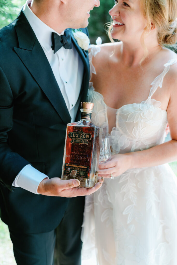 Groom holds dirty bottle of whiskey with bride holding a glass. 