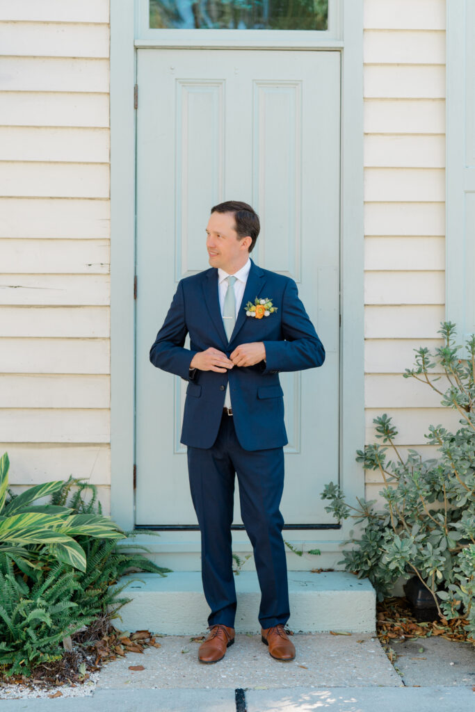 Groom in blue suit and light green tie buttons his jacket. 