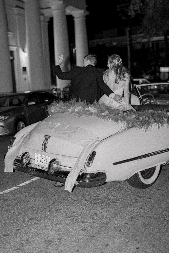 Bride and groom sit in the back of convertible getaway car and wave at the crowd as they walk away. 