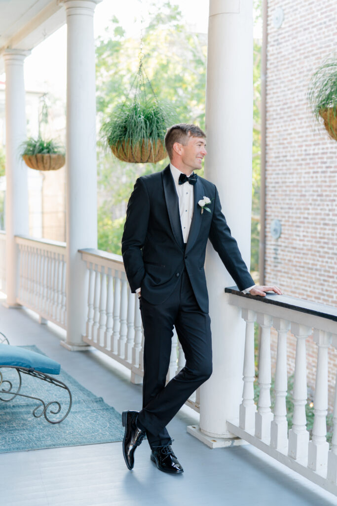 Groom in tuxedo leaning on porch railing of historic home in downtown Charleston wearing brackish bowtie. 