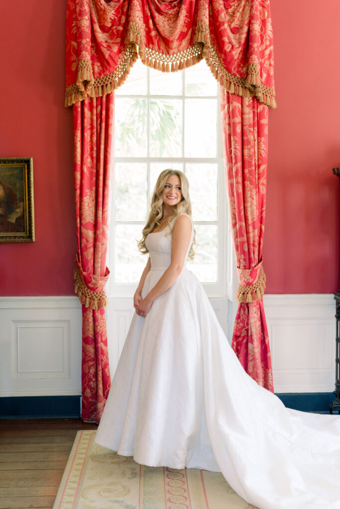 Bride puts on her wedding dress for the first time on wedding day in red room with red and gold curtains. 
