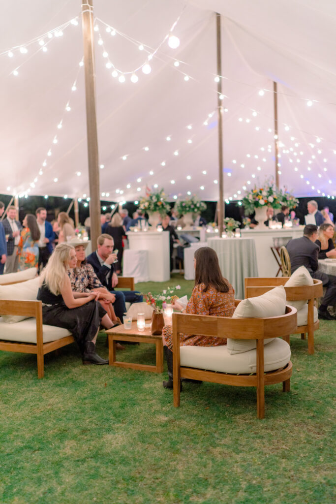 Wedding guests sit on furniture under sailcloth tent with string lights. Outdoor wedding welcome party in Charleston. 