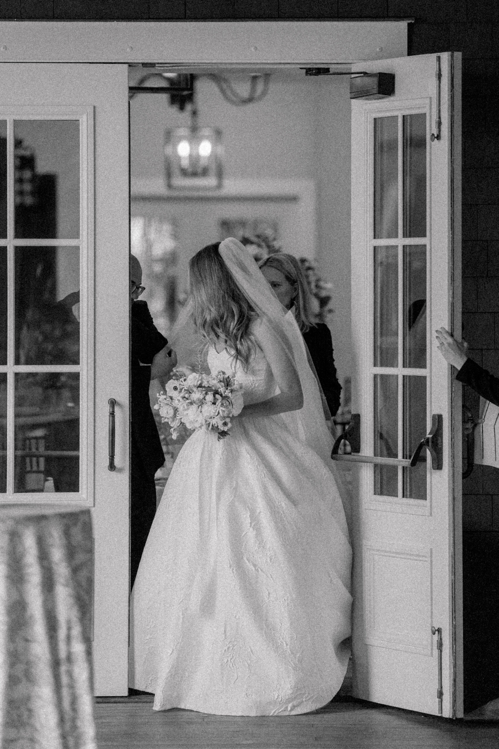 Candid black and white pre-wedding ceremony moment of bride trying to get her veil through a door. 