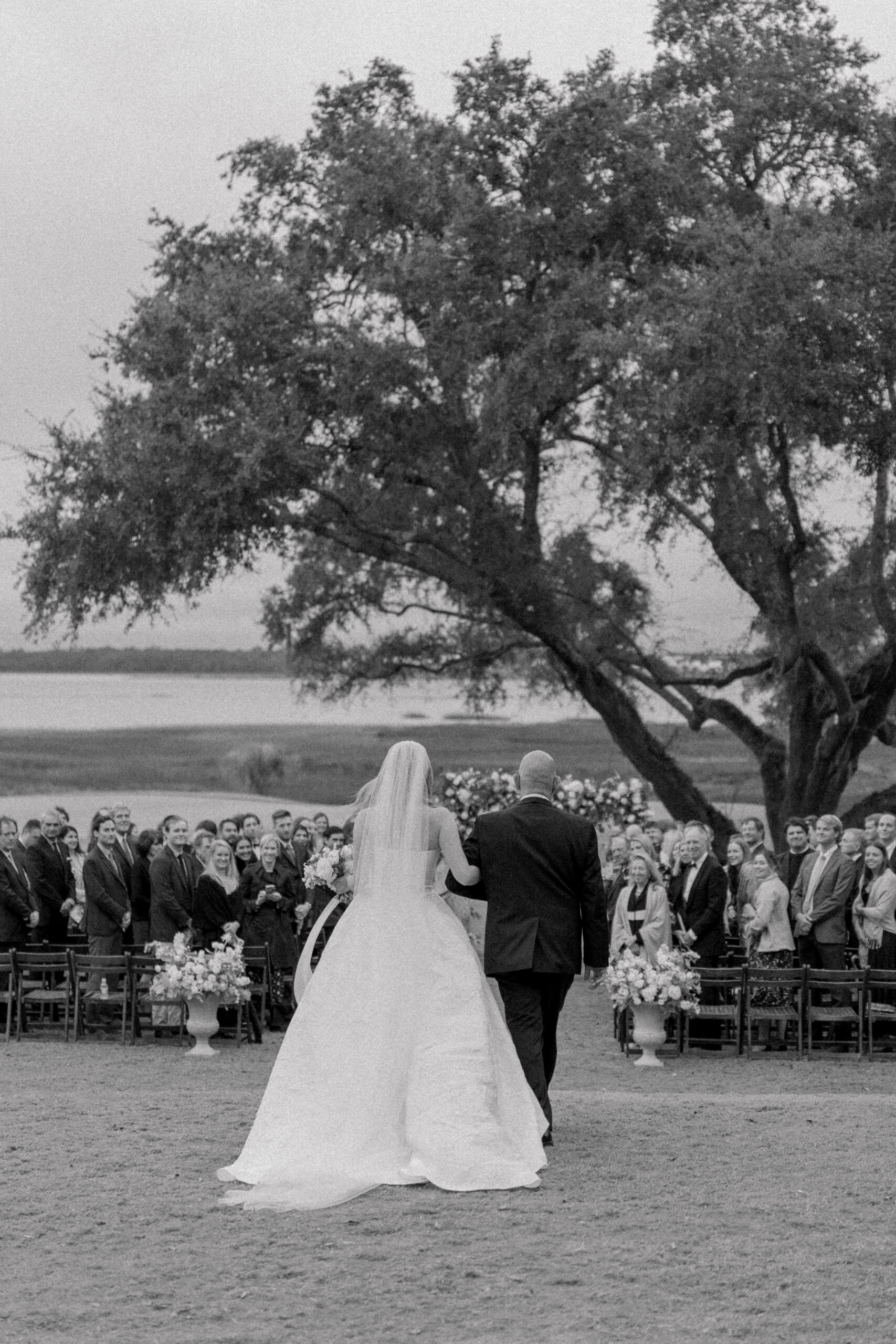 Father of the bride walks daughter up the aisle at waterfront wedding ceremony in Charleston. Black and white wedding photo. Live oak tree. 