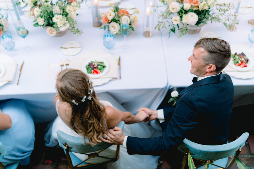 Bride and groom holding hands under the table and smiling during wedding day speeches. 