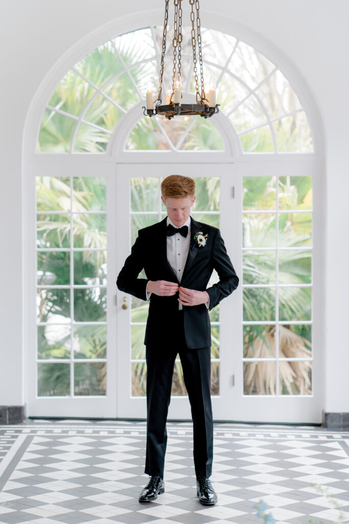 Groom in black tuxedo buttoning jacket with palm tree leaves in background and candle chandelier 