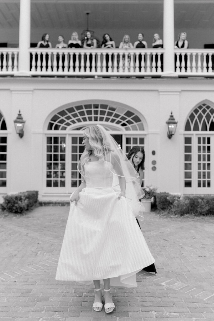 Black and white candid photo of bride walking in front of Lowndes Grove getting help from maid of honor with dress. 